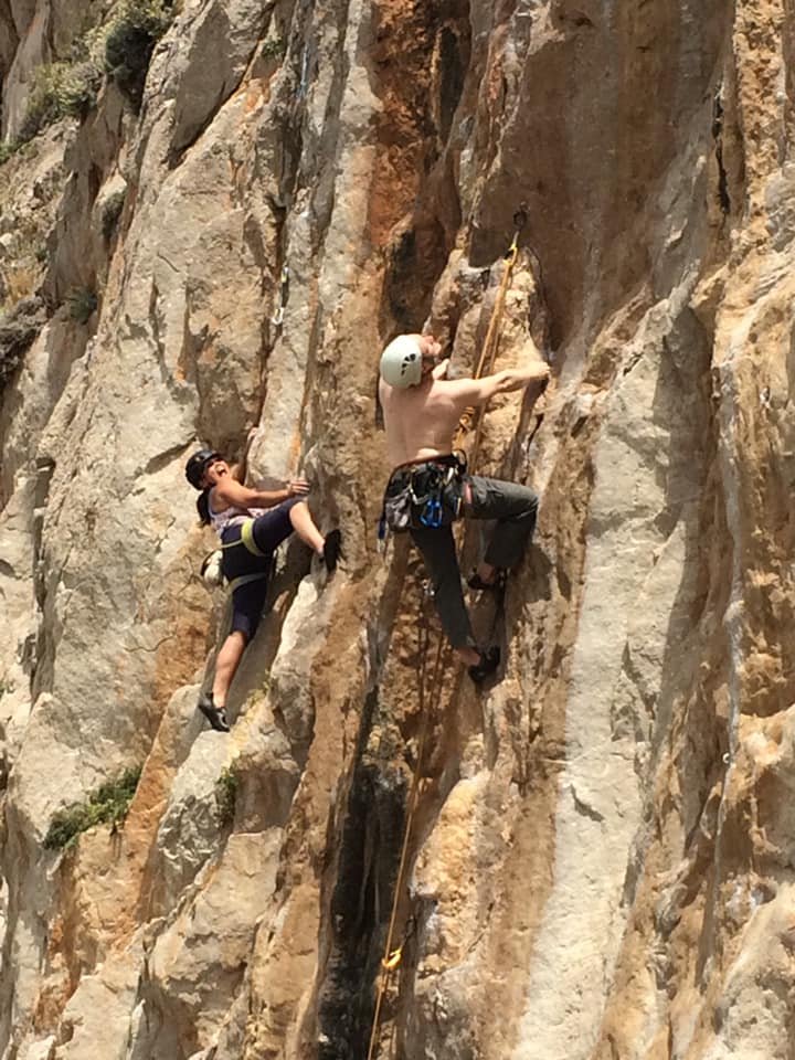 Two rock climbers, one male and one female, climbing a steep cliff face. The male climber is shirtless and wearing a helmet, while the female is wearing sunglasses and a harness.