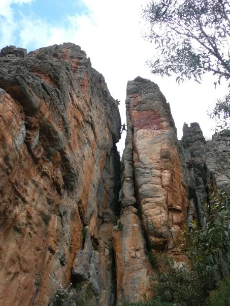 Tall, rugged rock formations with vertical cracks, with trees and foliage at the base, under a partly cloudy sky.