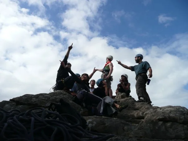 A group of people on a rocky hillside, some sitting and some standing, one person pointing upward and others gesturing, with climbing ropes in the foreground and a partly cloudy blue sky above.