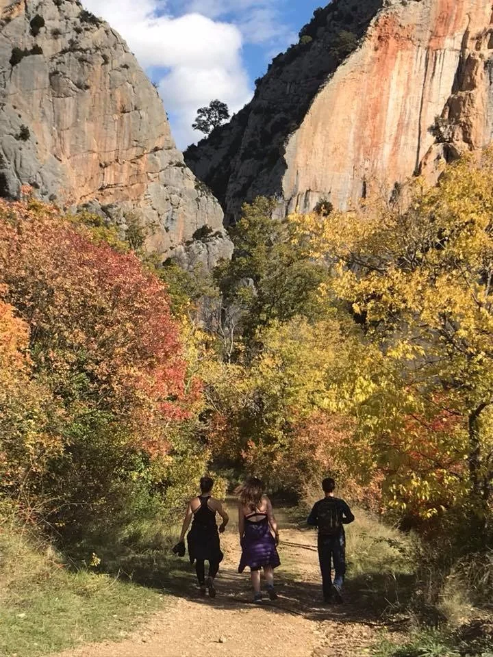 Three people walk along a dirt trail through a colorful autumn forest with tall cliffs in the background.