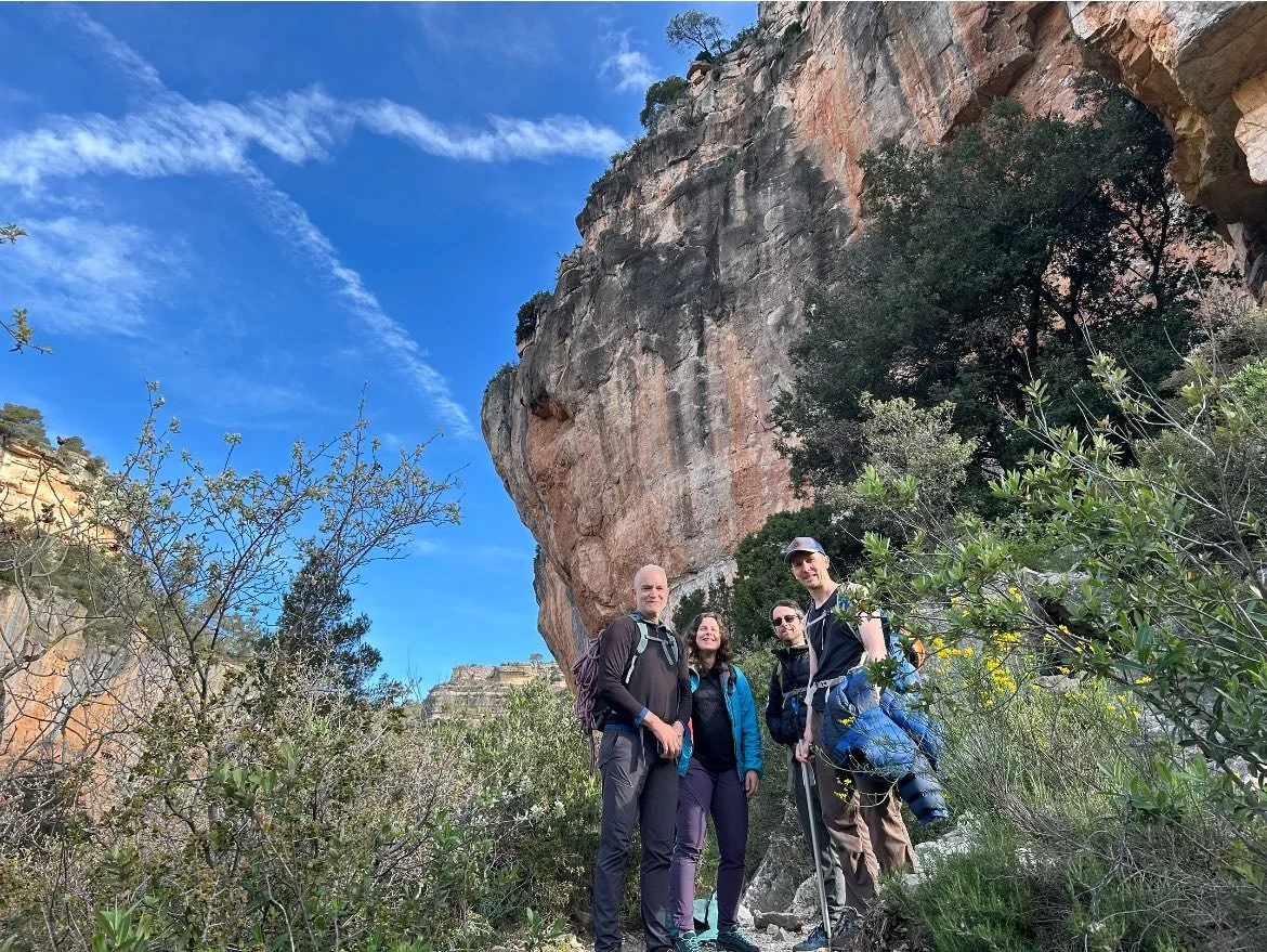 Four people hiking in a canyon with a large rocky cliffside and blue sky overhead.