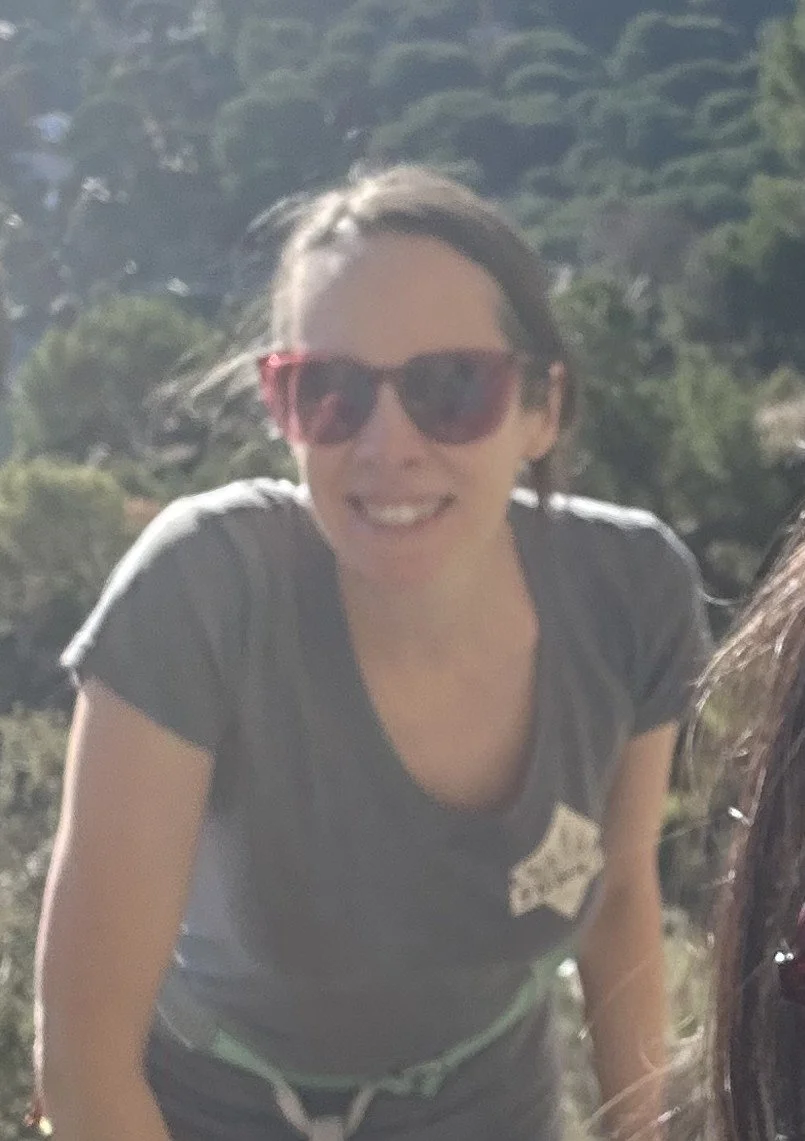 A smiling woman with dark hair wearing sunglasses and a gray T-shirt outdoors with trees in the background.