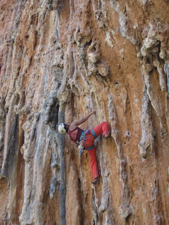 A person rock climbing on a steep, textured orange-brown cliff, wearing a helmet, harness, and climbing gear.