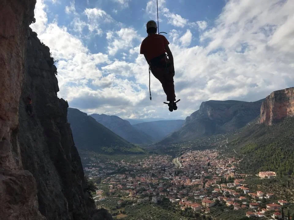 A person wearing a helmet is hanging upside down from a cliff, overlooking a town in a valley surrounded by mountains under a cloudy sky.