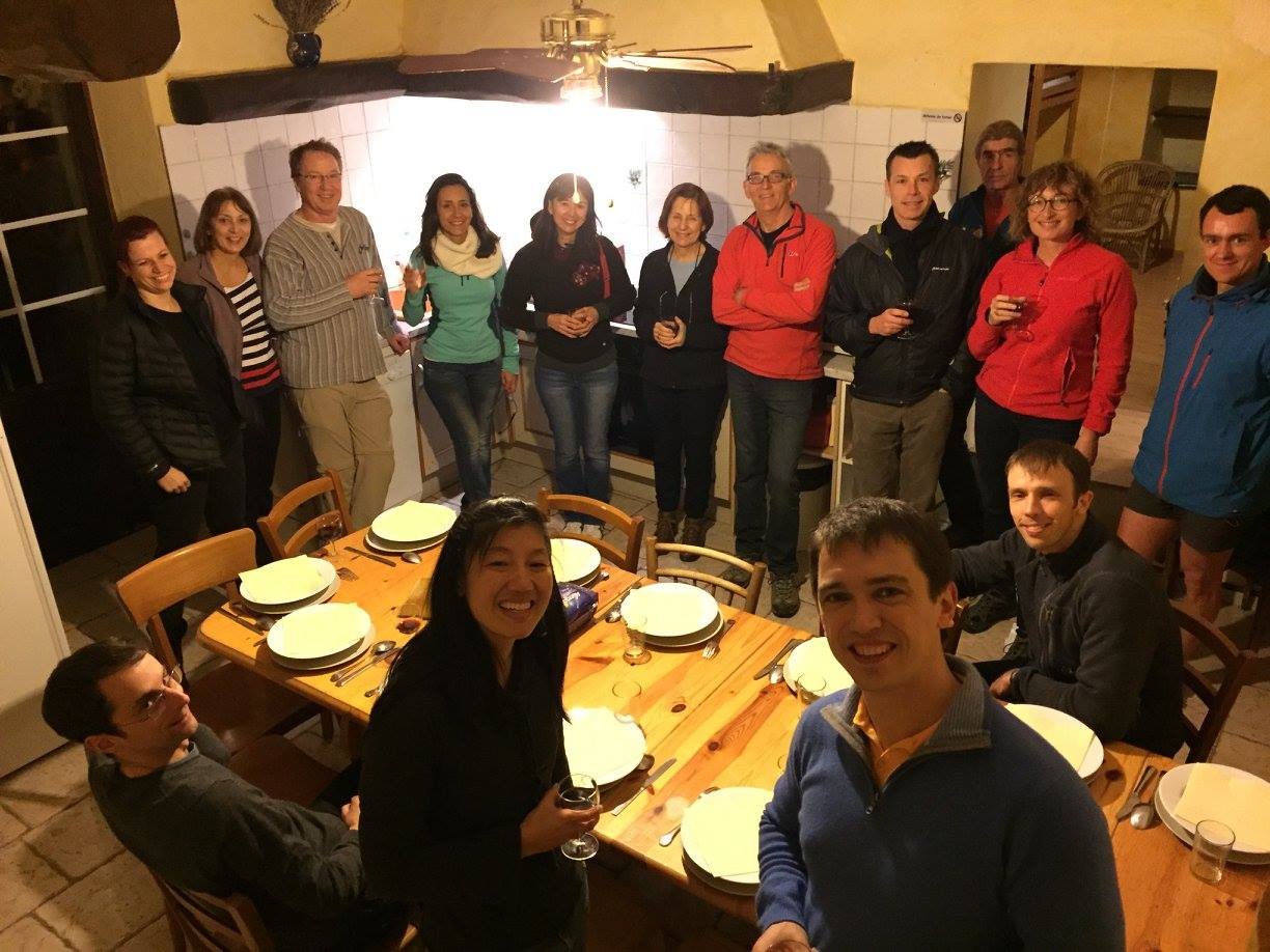 Group of people celebrating around a dining table in a cozy kitchen or dining area, some holding glasses of wine, with a large wooden table set with dishes and cutlery, and a ceiling fan overhead.