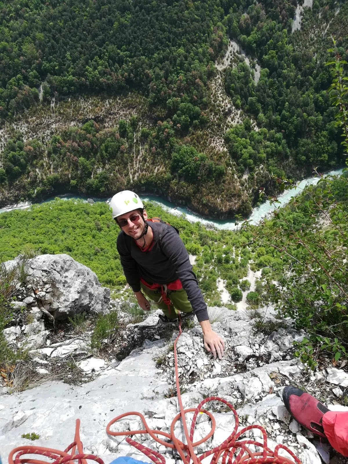 A man wearing a helmet and sunglasses rock climbing on a steep, rocky mountain face with a forested landscape and river below in the background.