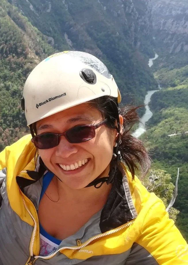 Woman in yellow and gray jacket wearing a white helmet and sunglasses, smiling with a scenic river and green valley in the background.