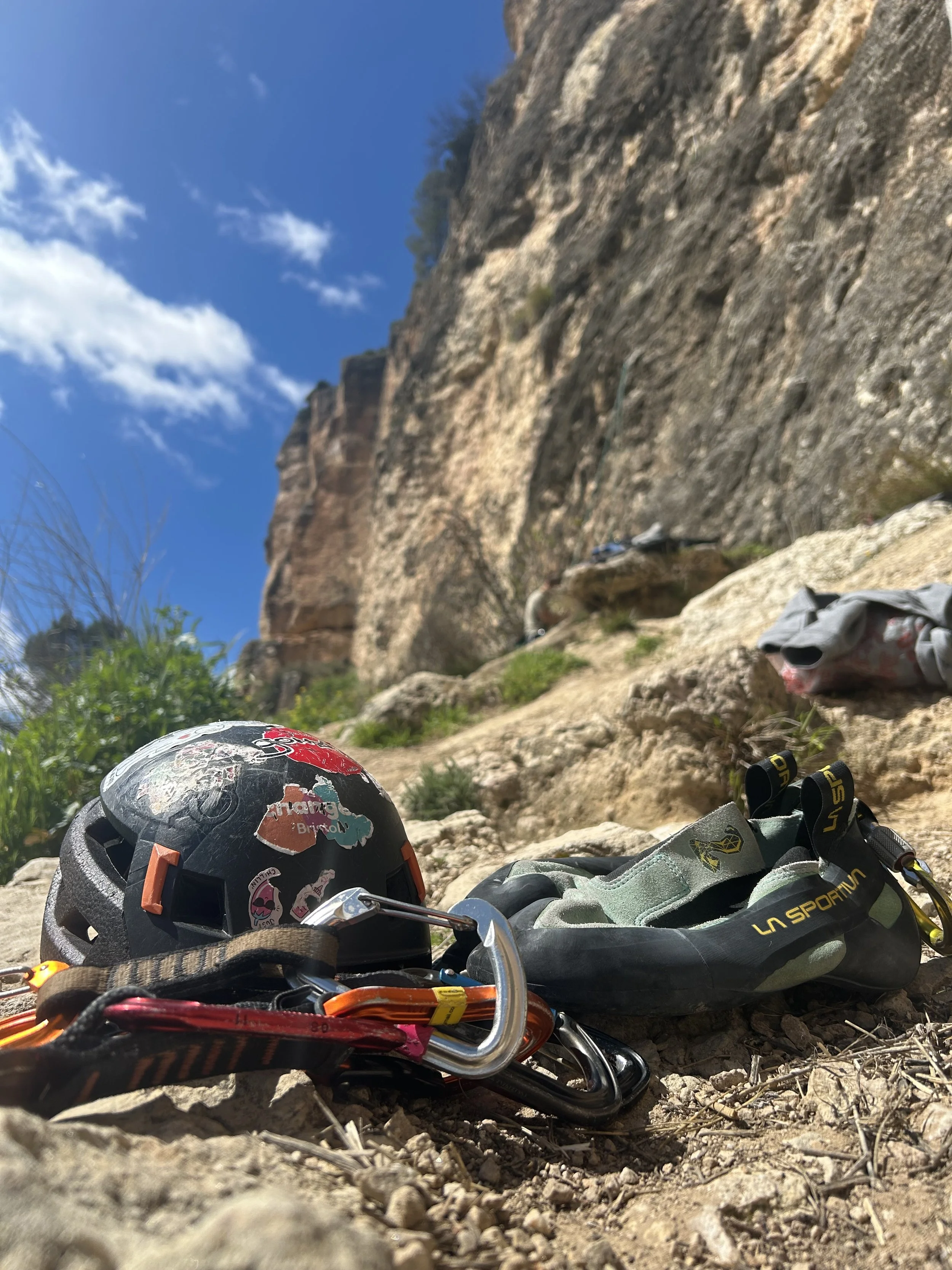 Climbing gear, including a helmet with stickers, carabiners, and climbing shoes, placed on rocky ground with a mountain cliff and blue sky with clouds in the background.