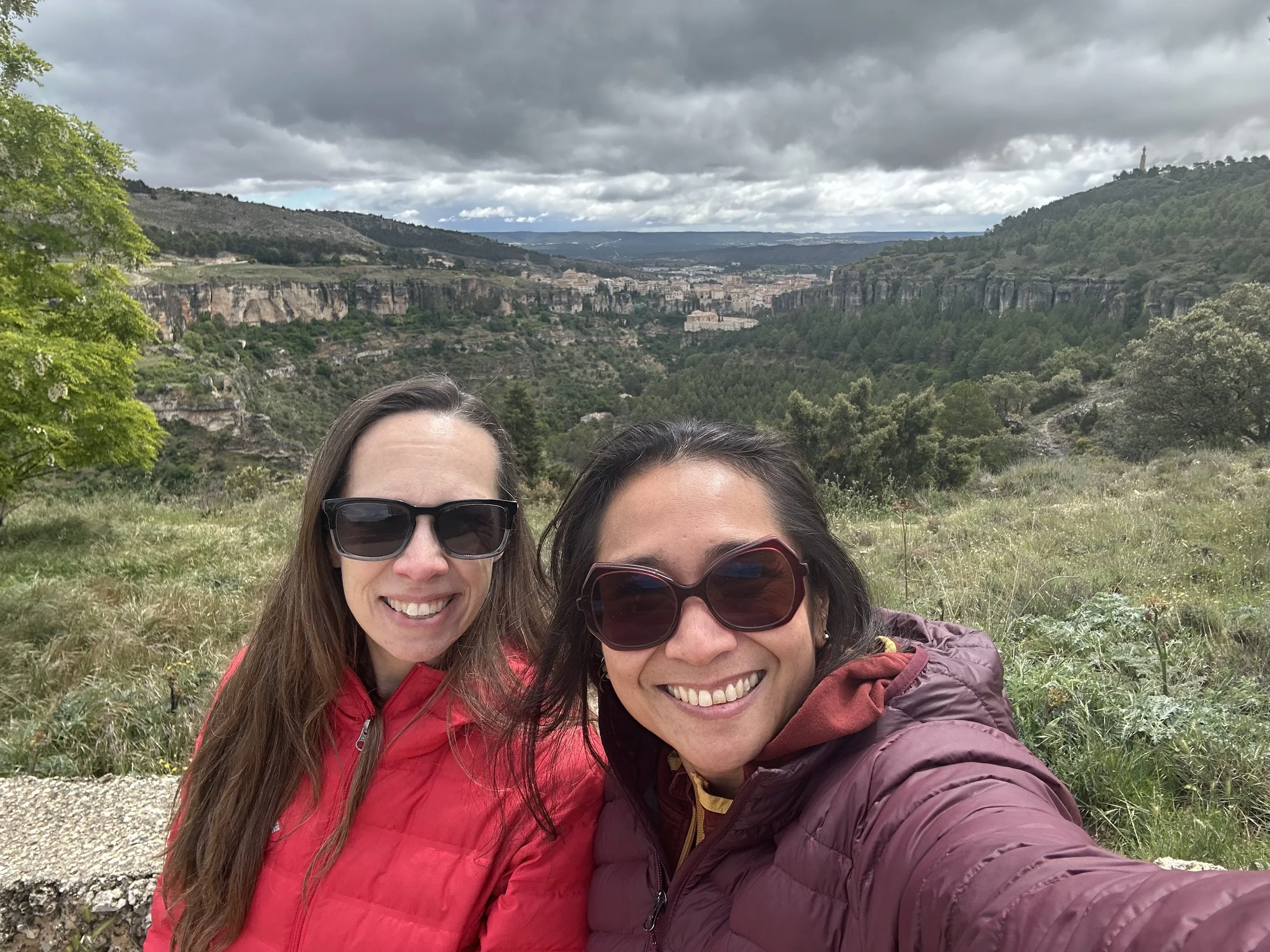 Two women smiling and wearing sunglasses taking a selfie outdoors with a hilly landscape and cloudy sky in the background.
