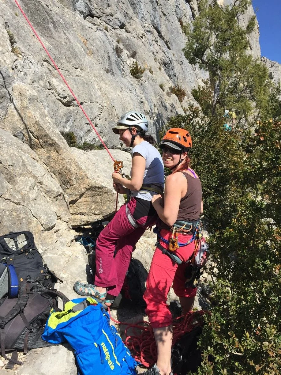Two women wearing helmets rock climbing on a rocky cliff, holding climbing ropes, with climbing gear and backpacks on the ground