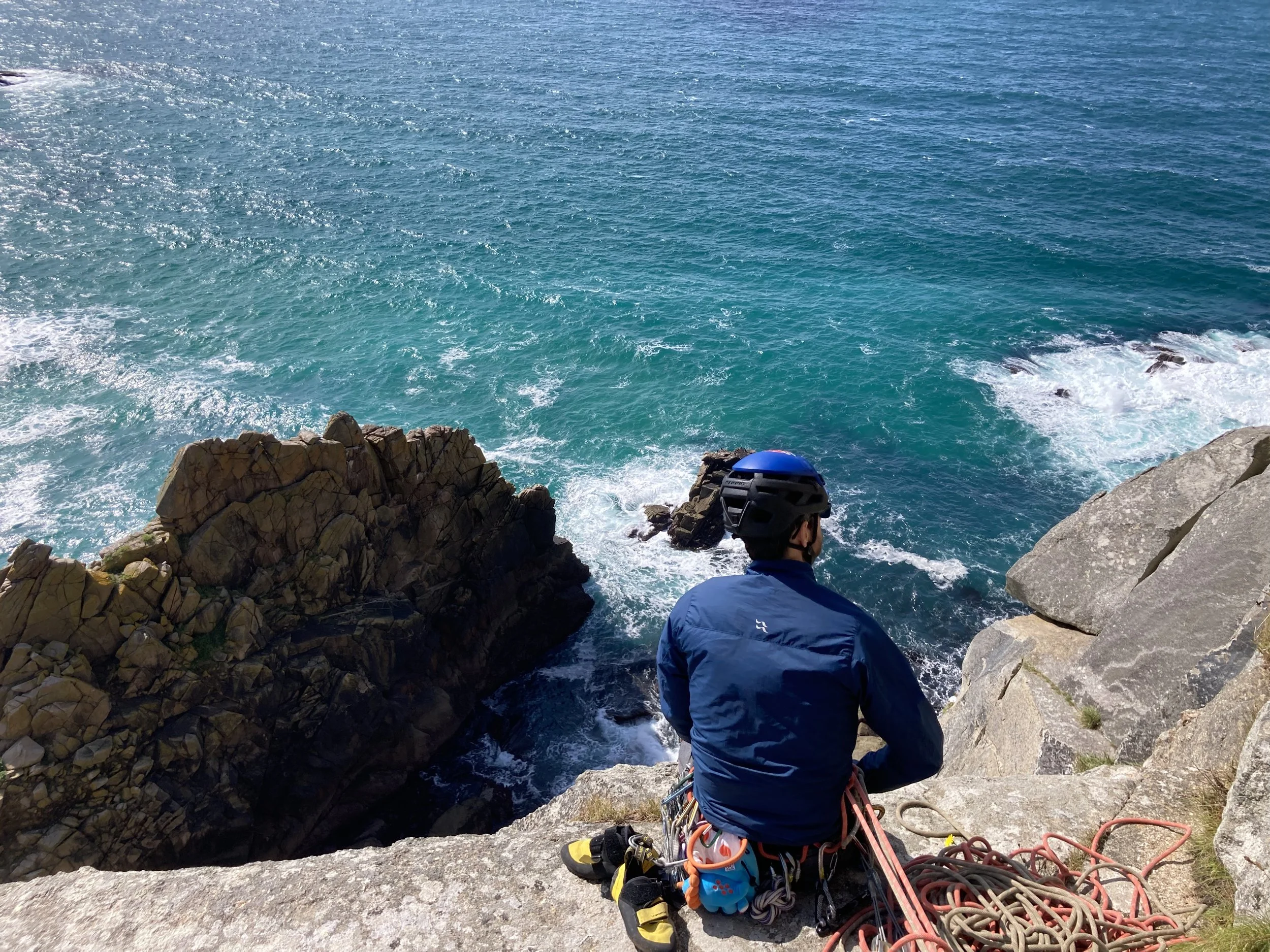 A person with climbing gear sitting on a rocky ledge overlooking the ocean, with the sea and rocks visible below.