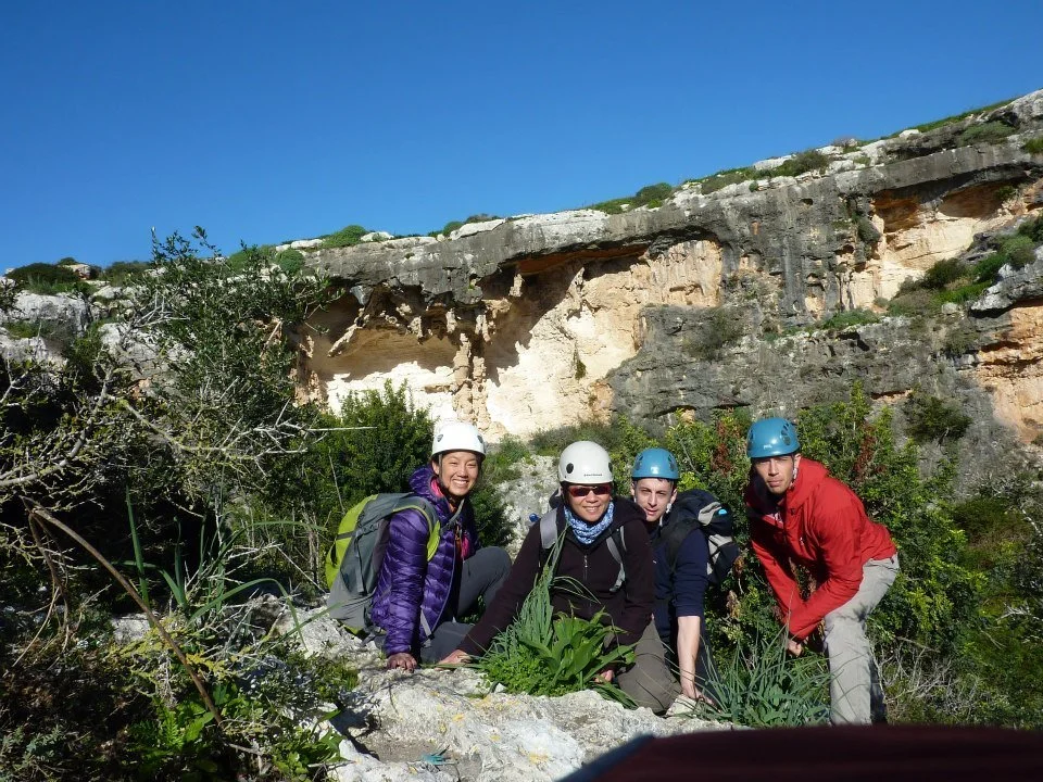 Four people wearing helmets and outdoor gear, sitting and squatting on rocky terrain with green plants, in front of a cliff and blue sky.