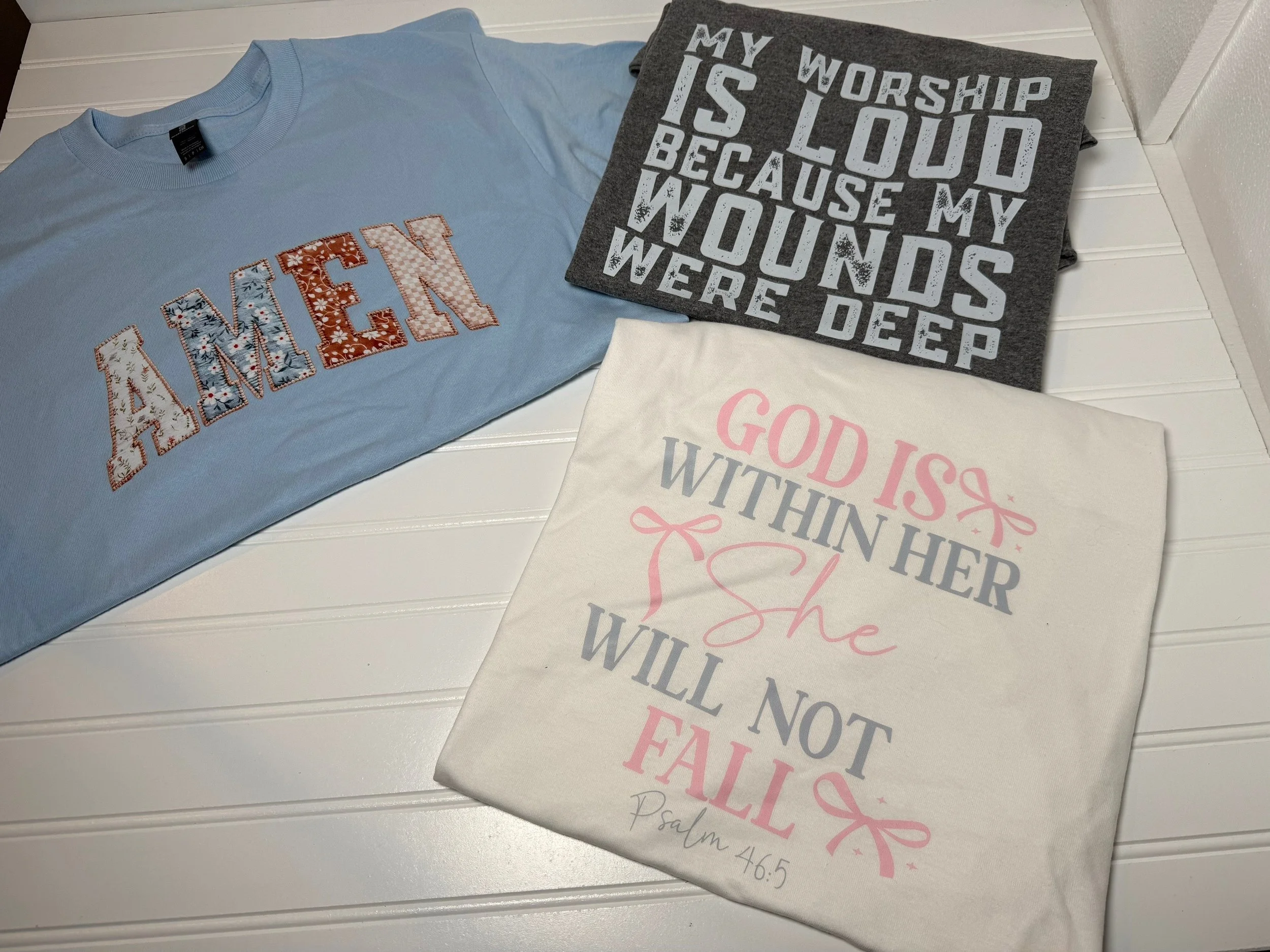 Three T-shirts with religious and motivational messages laid out on a white wooden surface.