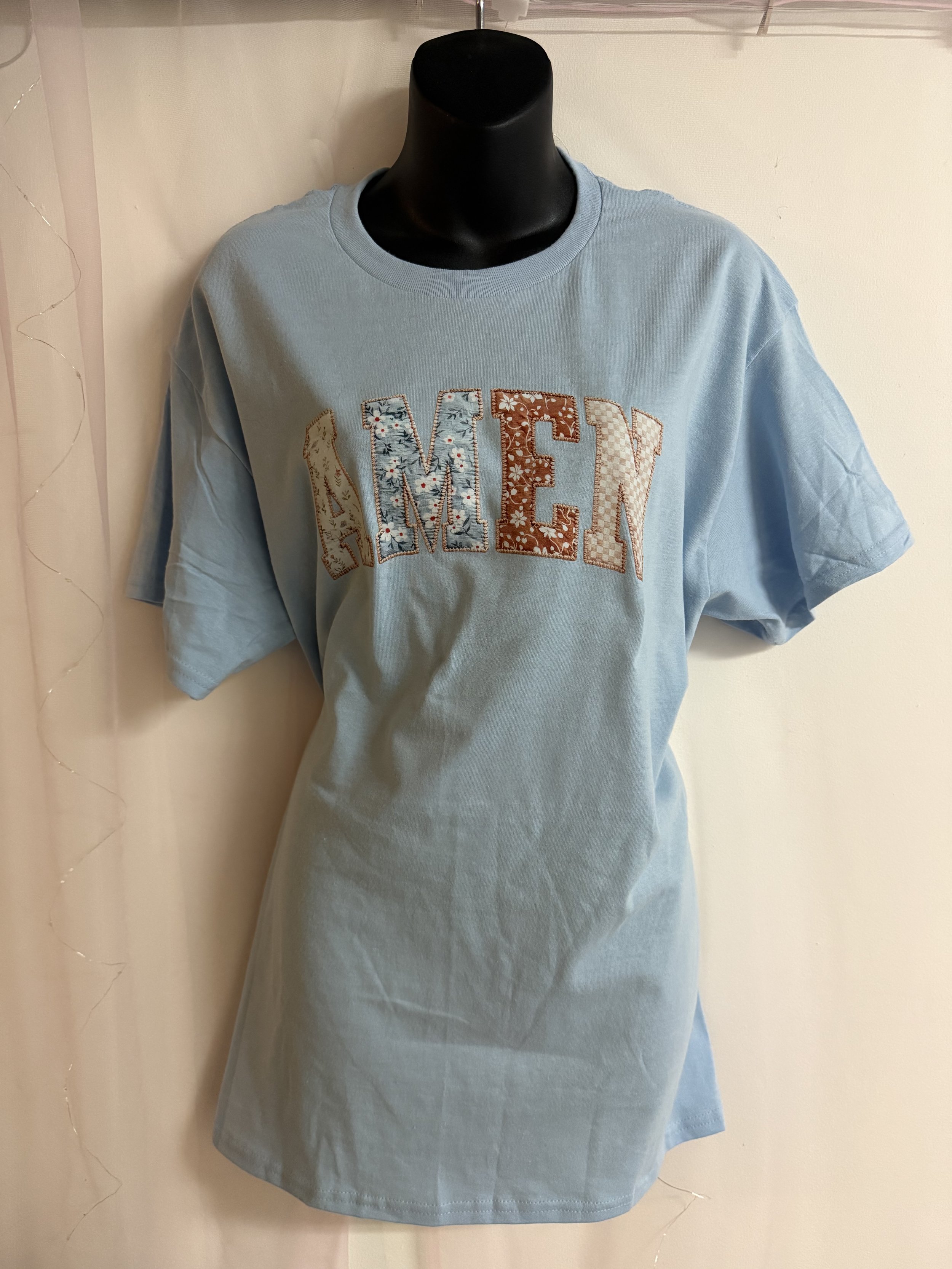 Light blue T-shirt on a mannequin with embroidered patch letters spelling 'NEW' in floral and checkered designs.