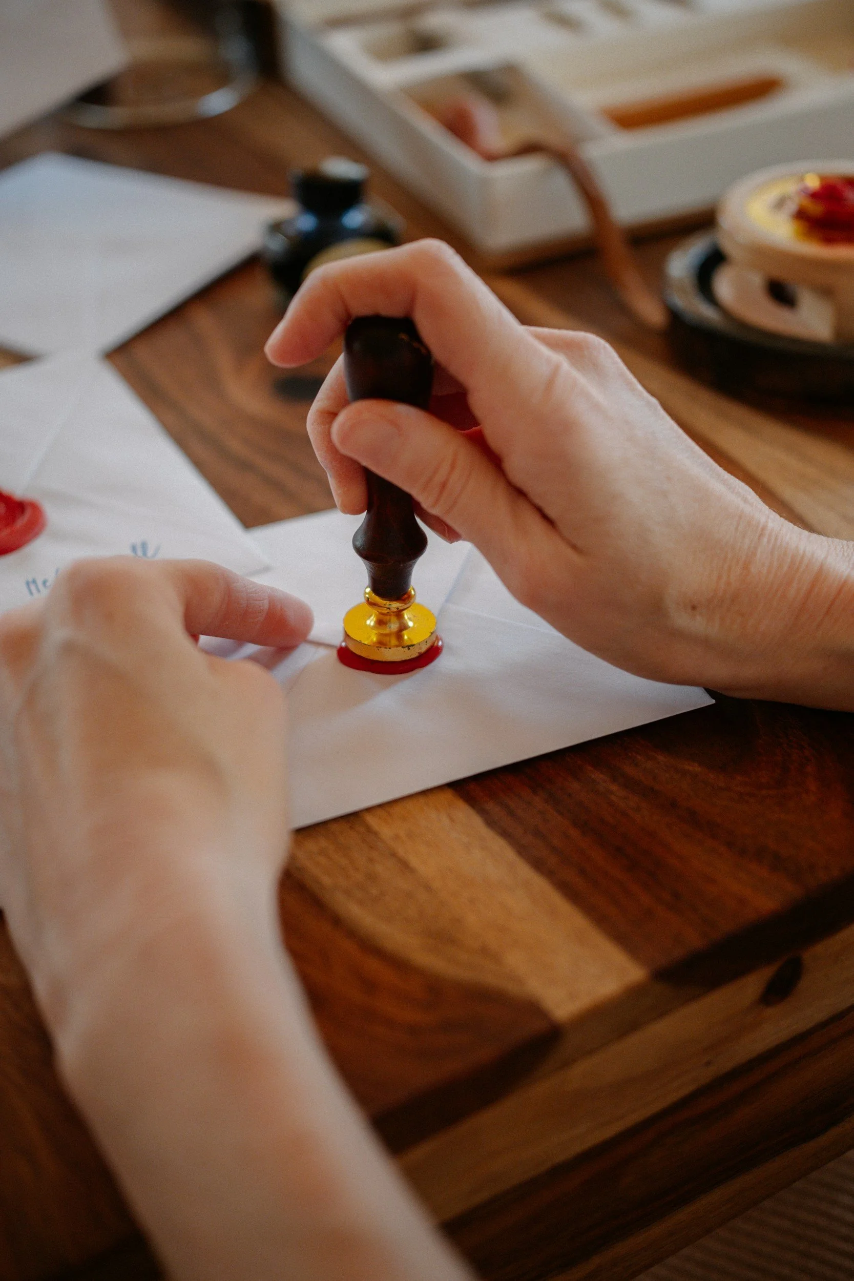 Person holding a black and gold wax seal stamp over a sheet of paper, with a wooden desk, papers, and a printer in the background.