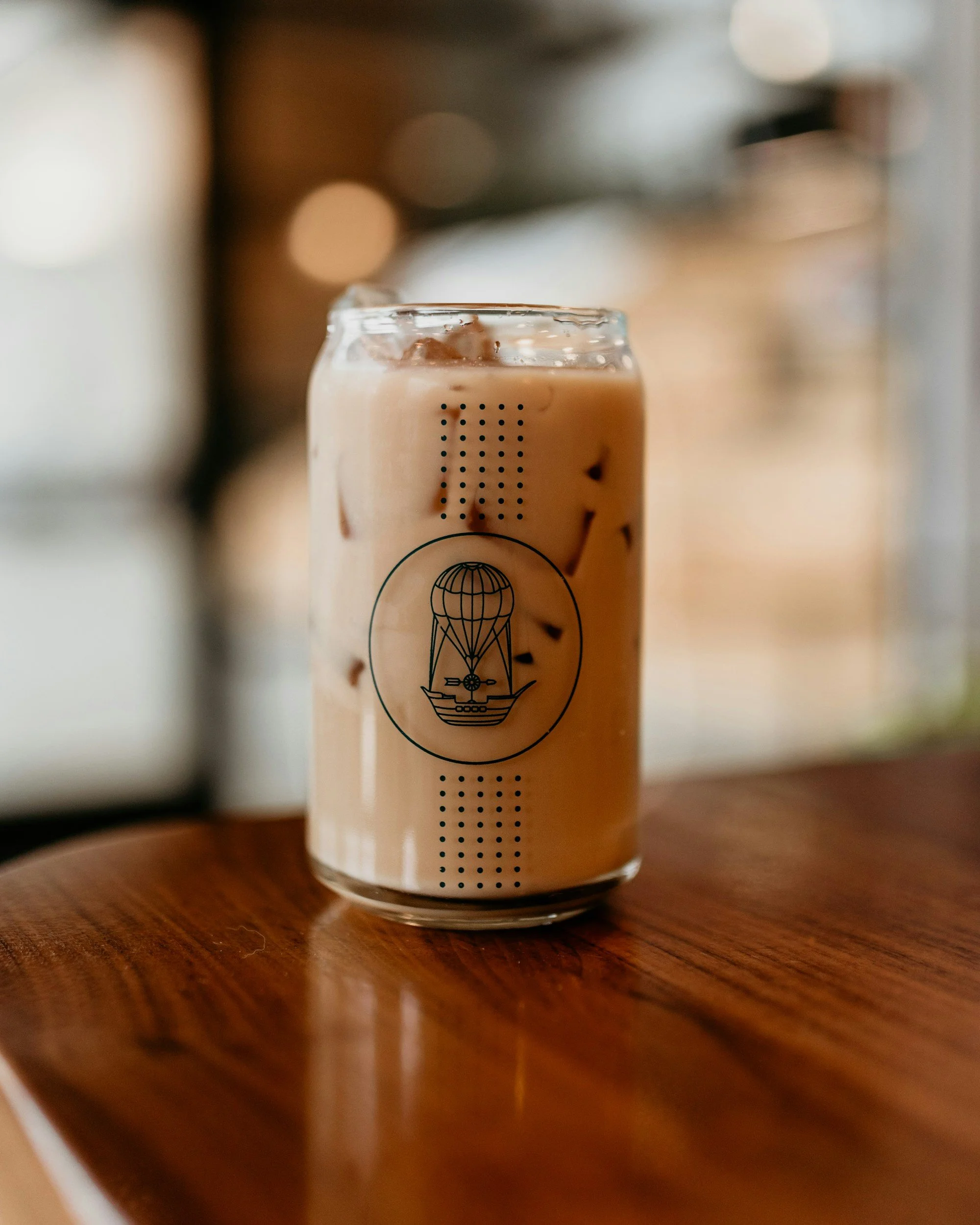 Close-up of an iced coffee drink in a clear glass with a logo, placed on a wooden surface, with a blurred background.