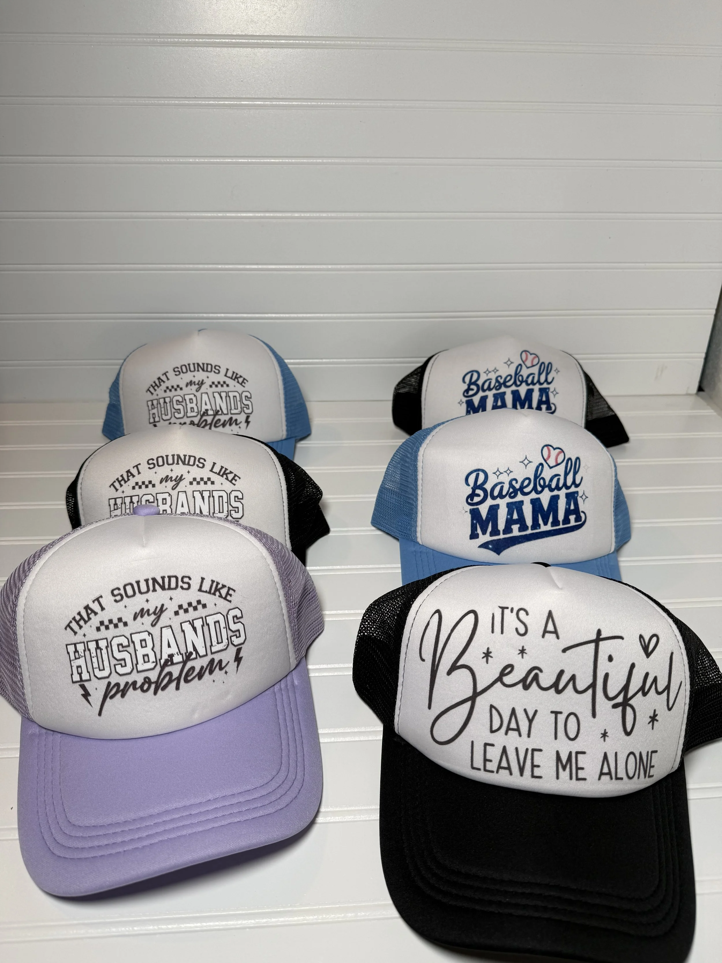 Multiple trucker hats displayed on a white surface, with one featuring the text 'That sounds like my husband problem!' and another saying 'Baseball Mama'.