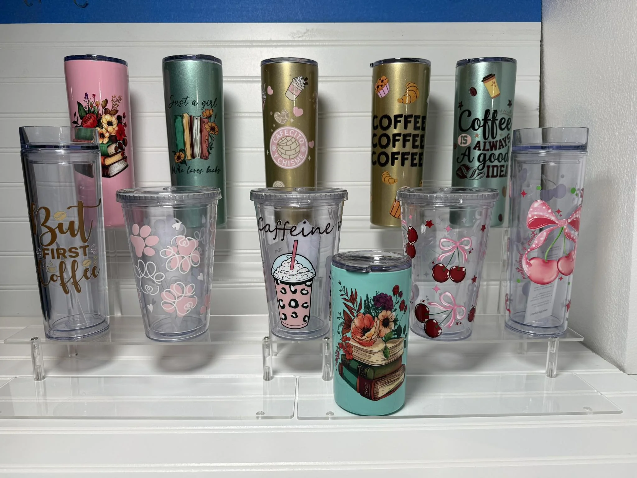 Display of colorful travel tumblers with various designs, including books, flowers, cupcakes, coffee cups, and cherries, arranged on a clear shelf with empty clear plastic cups in front.