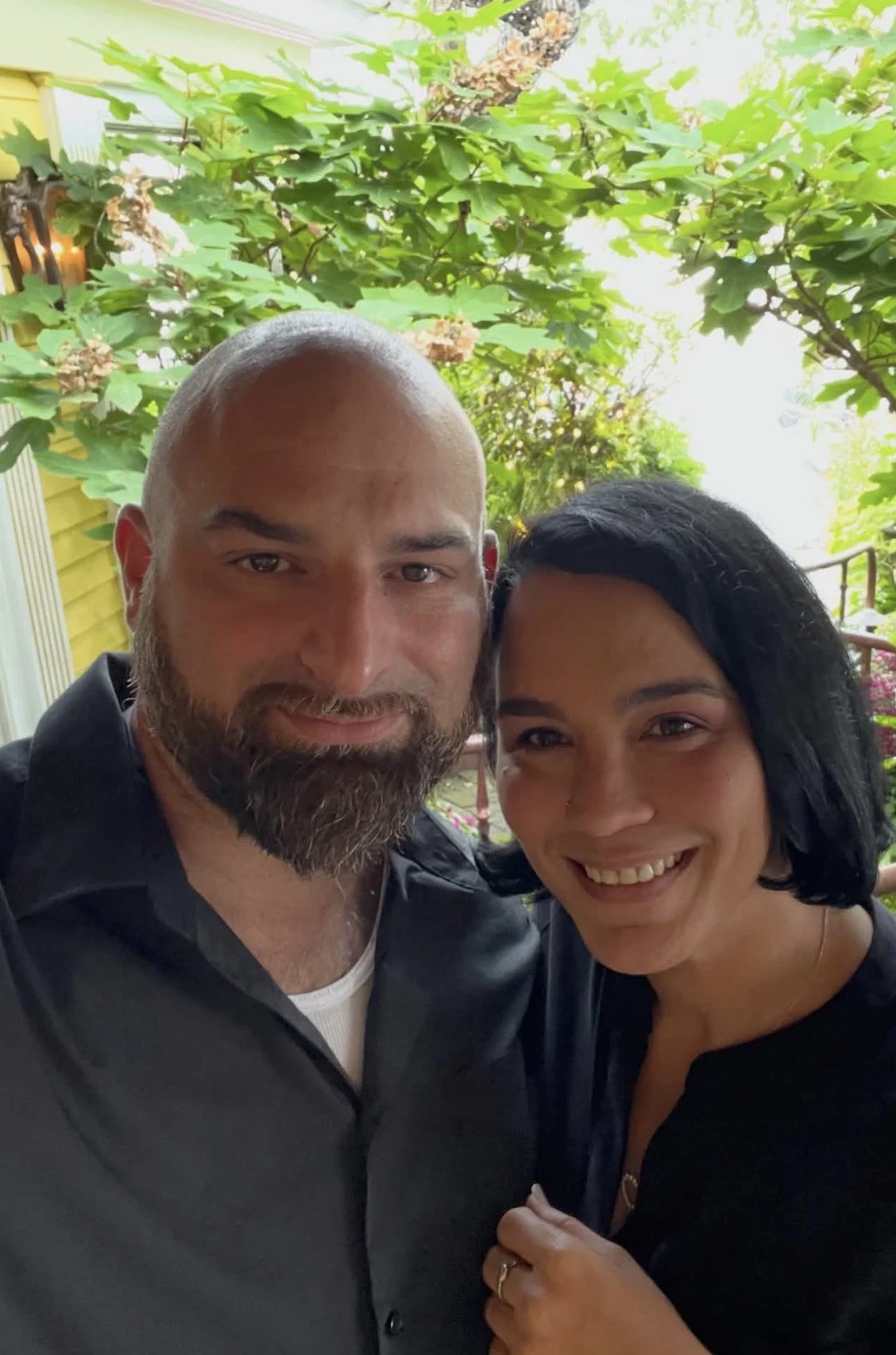 A smiling man with a beard and a woman with black hair, posing together outdoors in front of green leafy trees.