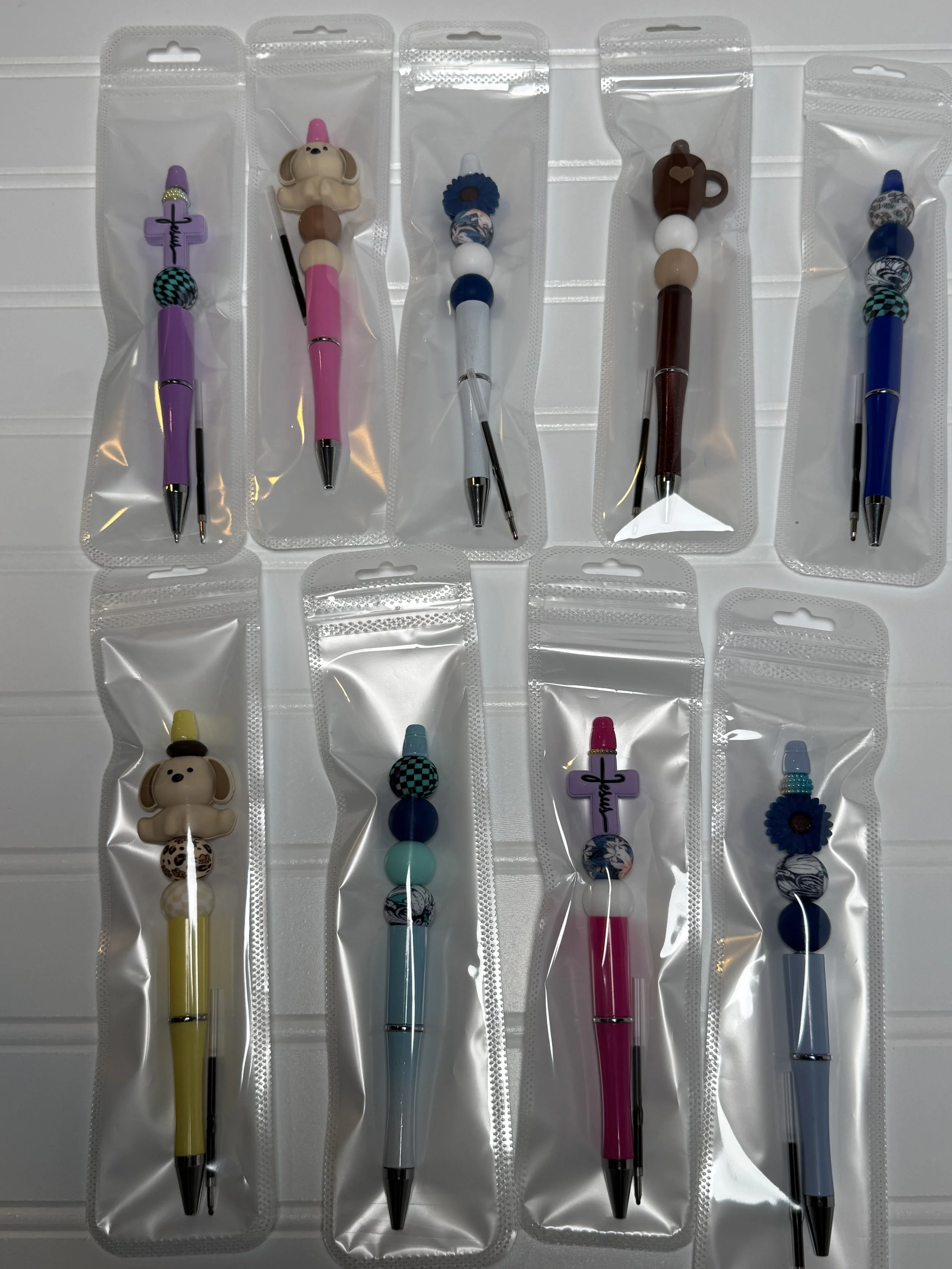 Colorful pens with decorative charms, in individual clear plastic packaging, lined up on a white surface.