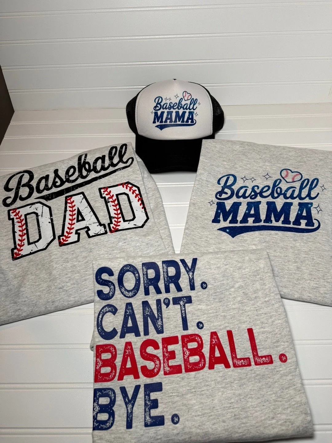 A baseball cap and three T-shirts with baseball-themed phrases, including "Baseball Mama", "Baseball Dad", and "Sorry, can't baseball bye", laid out on a white surface.
