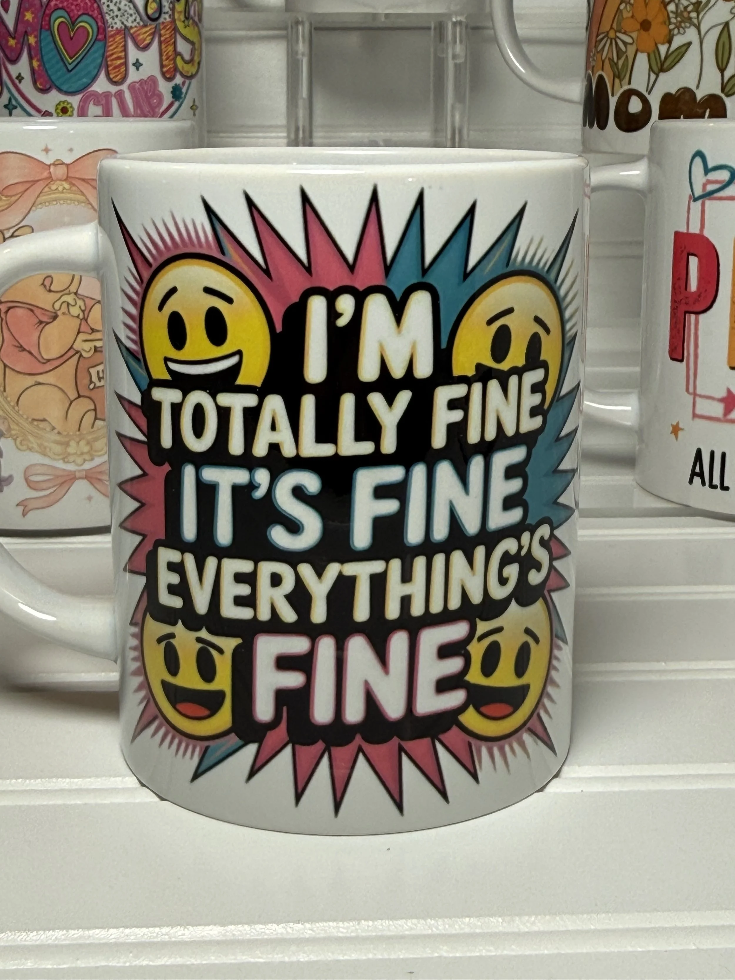 A white coffee mug with colorful comic-style text that reads "I'm totally fine, it's fine, everything's fine" accompanied by emoticon faces expressing mixed emotions. Other similar mugs are visible in the background.