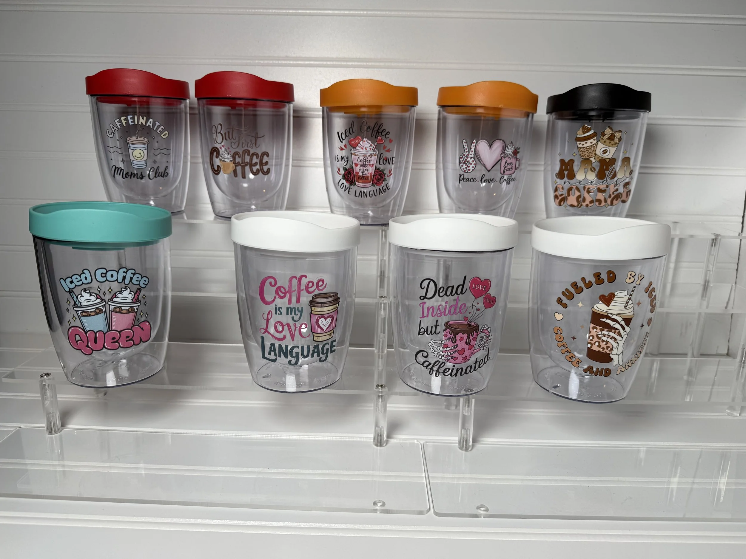 Collection of clear plastic tumblers with colorful lids and printed designs, arranged on a white shelf.