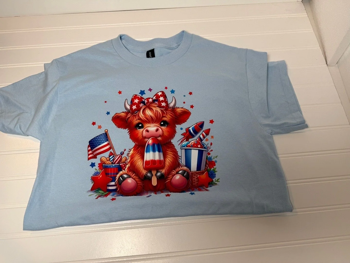 Light blue T-shirt featuring a cartoon of a cute pig with a red, white, and blue bow, surrounded by patriotic decorations like an American flag, rockets, stars, and a bucket of fireworks, with red and blue confetti.