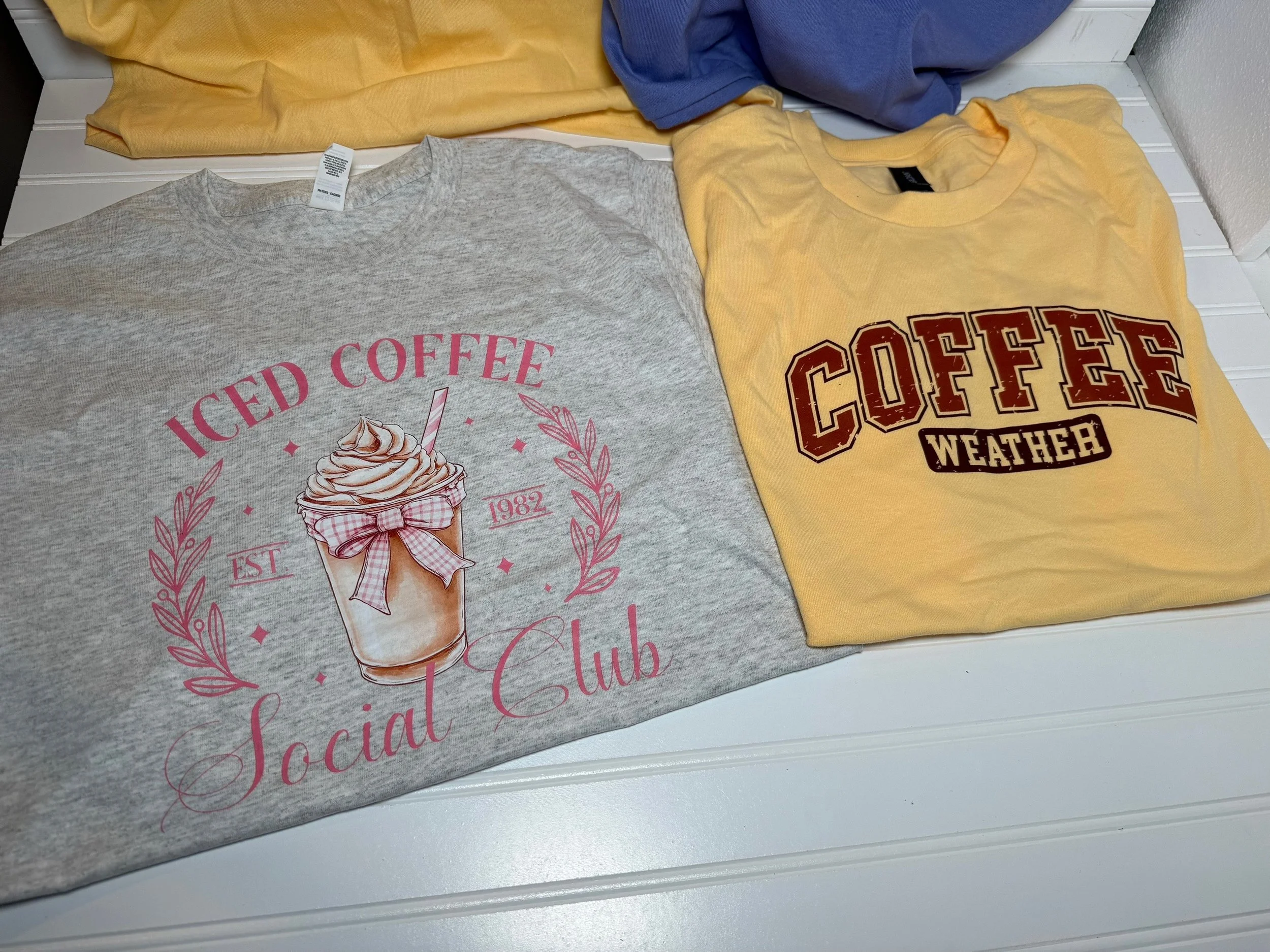 Two folded T-shirts, one gray with pink text and a graphic of an iced coffee drink, and the other yellow with a vintage style Coffee Weather logo, lying on a white surface.