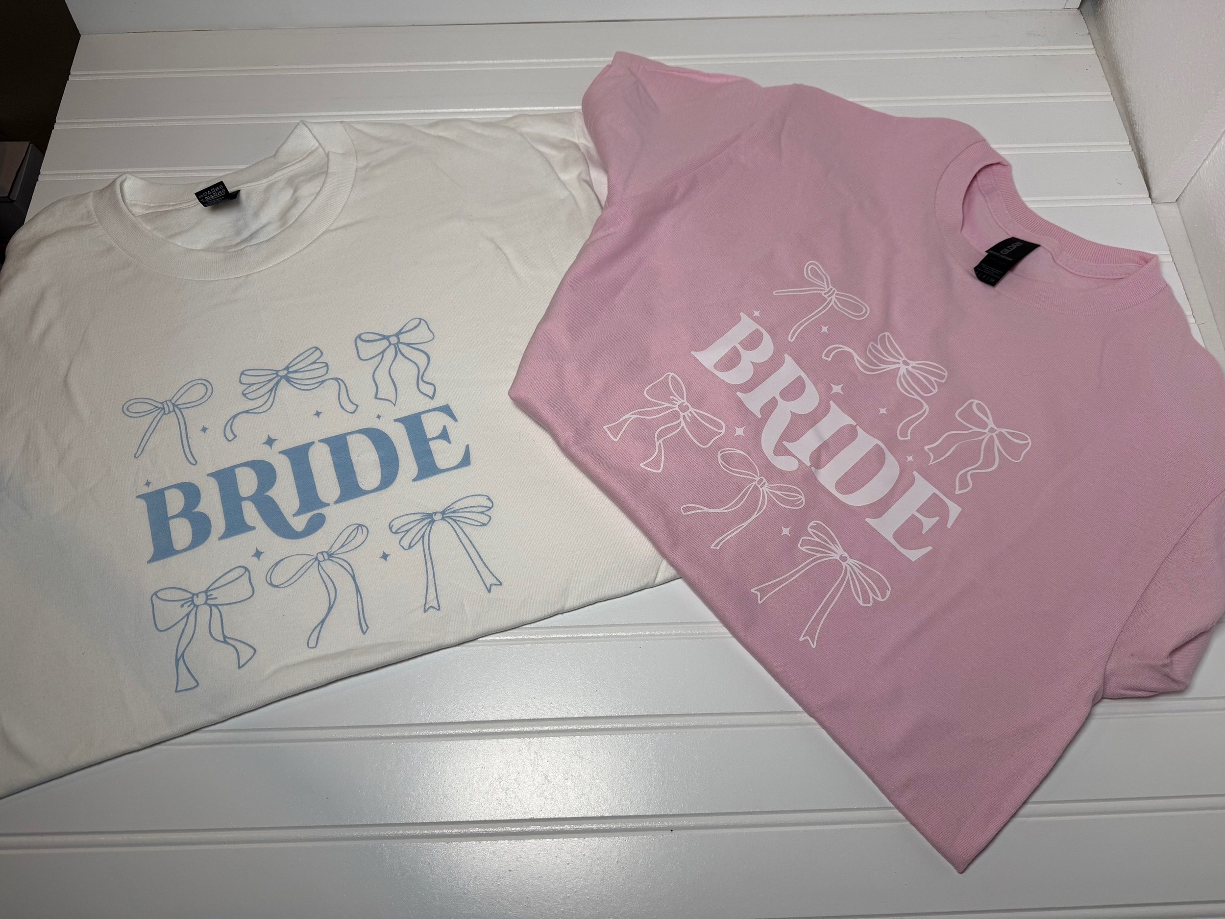 Two T-shirts laid flat on a white surface, one white with blue lettering and bows, and one pink with white lettering and bows.