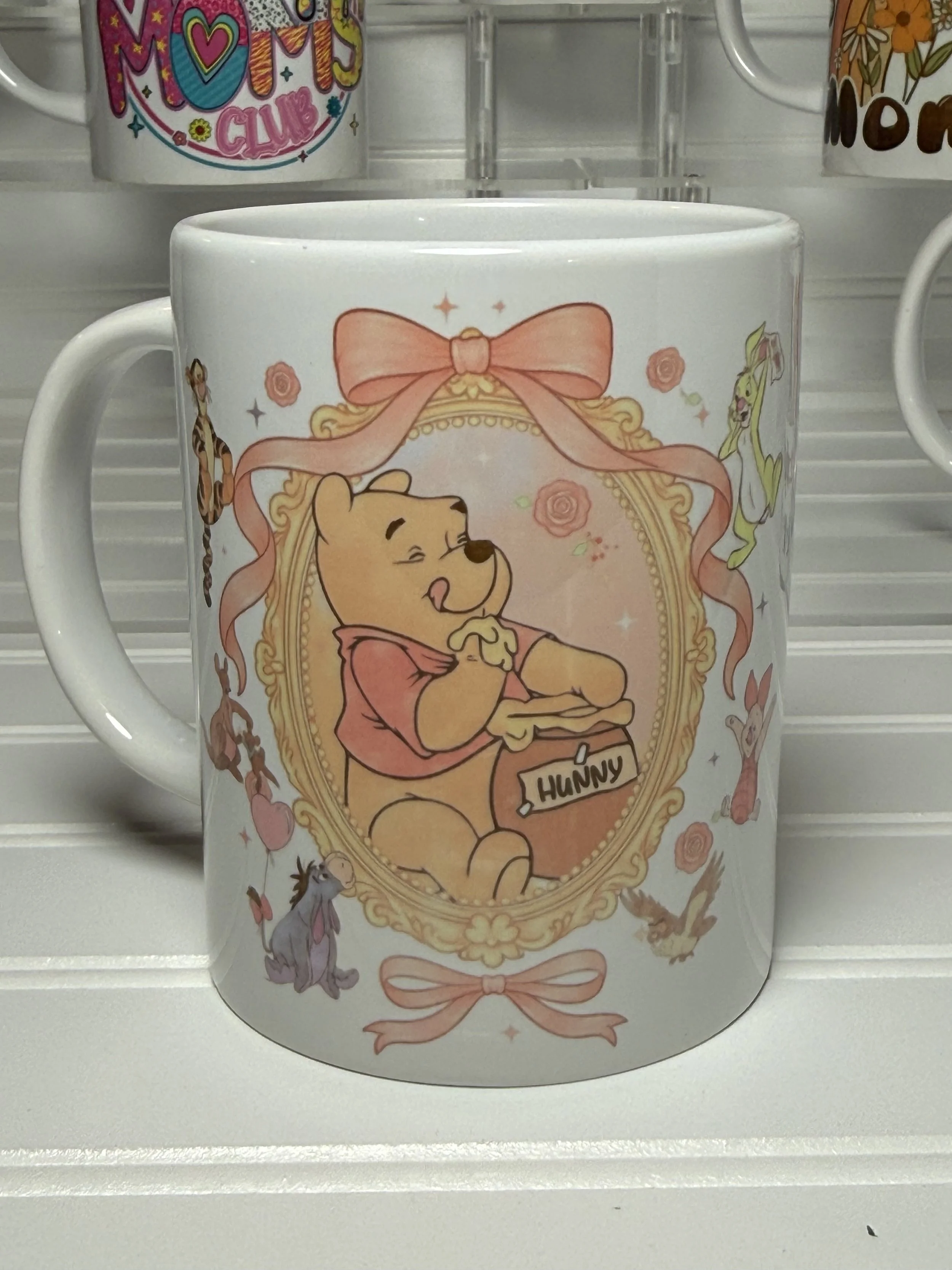 A white ceramic mug featuring Winnie the Pooh, who is smiling and holding a honey pot labeled 'HUNNY.' The mug is decorated with a pink bow, roses, and Disney characters including a giraffe and a rabbit.