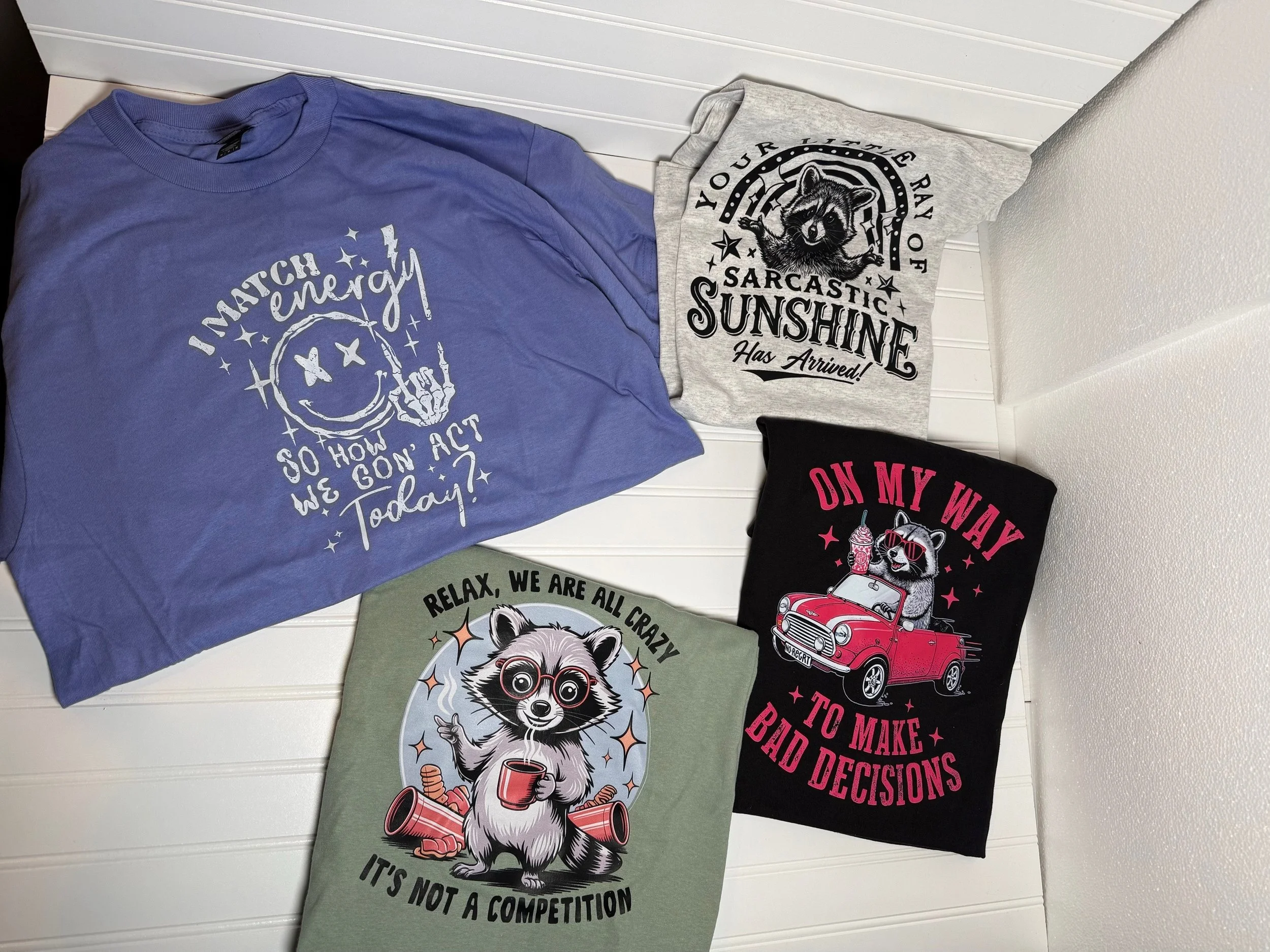 Collection of colorful graphic t-shirts with various humorous and motivational designs laid out on a white surface.