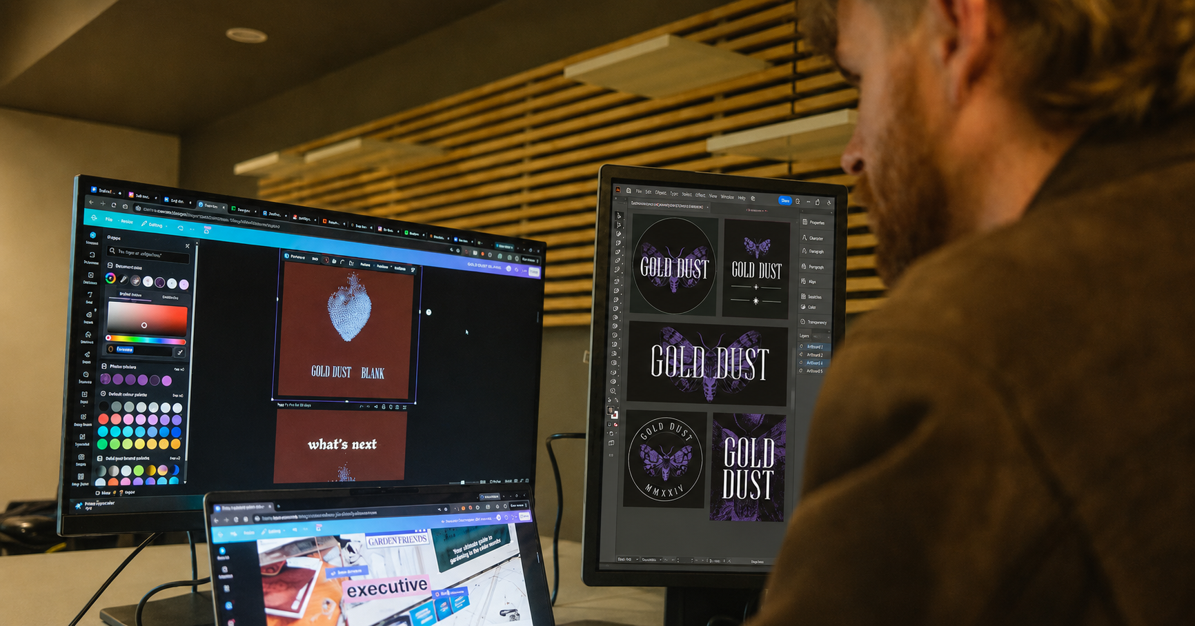 A person working on a computer with multiple monitors displaying graphic design programs and logos for 'Gold Dust', featuring a purple moth or butterfly design.