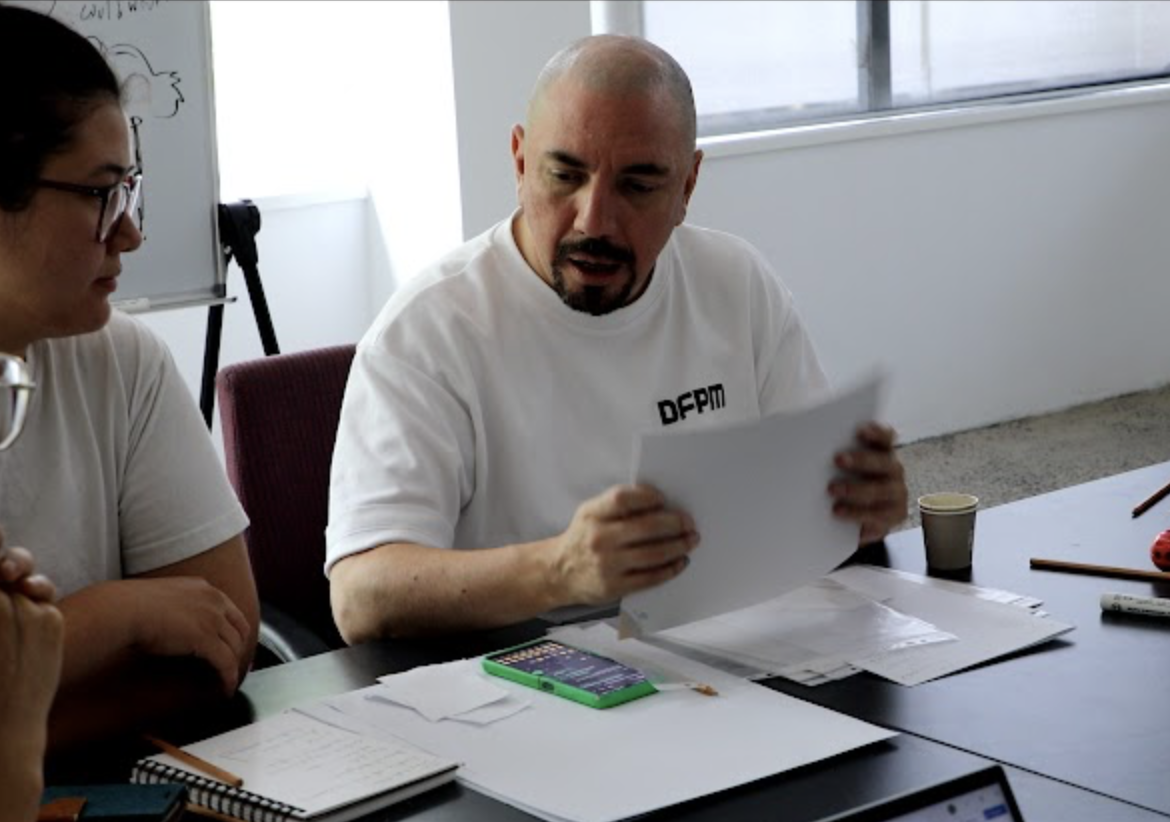 Two individuals sitting at a table during a discussion, with papers, a cell phone, and writing utensils on the table, in a bright room with windows.