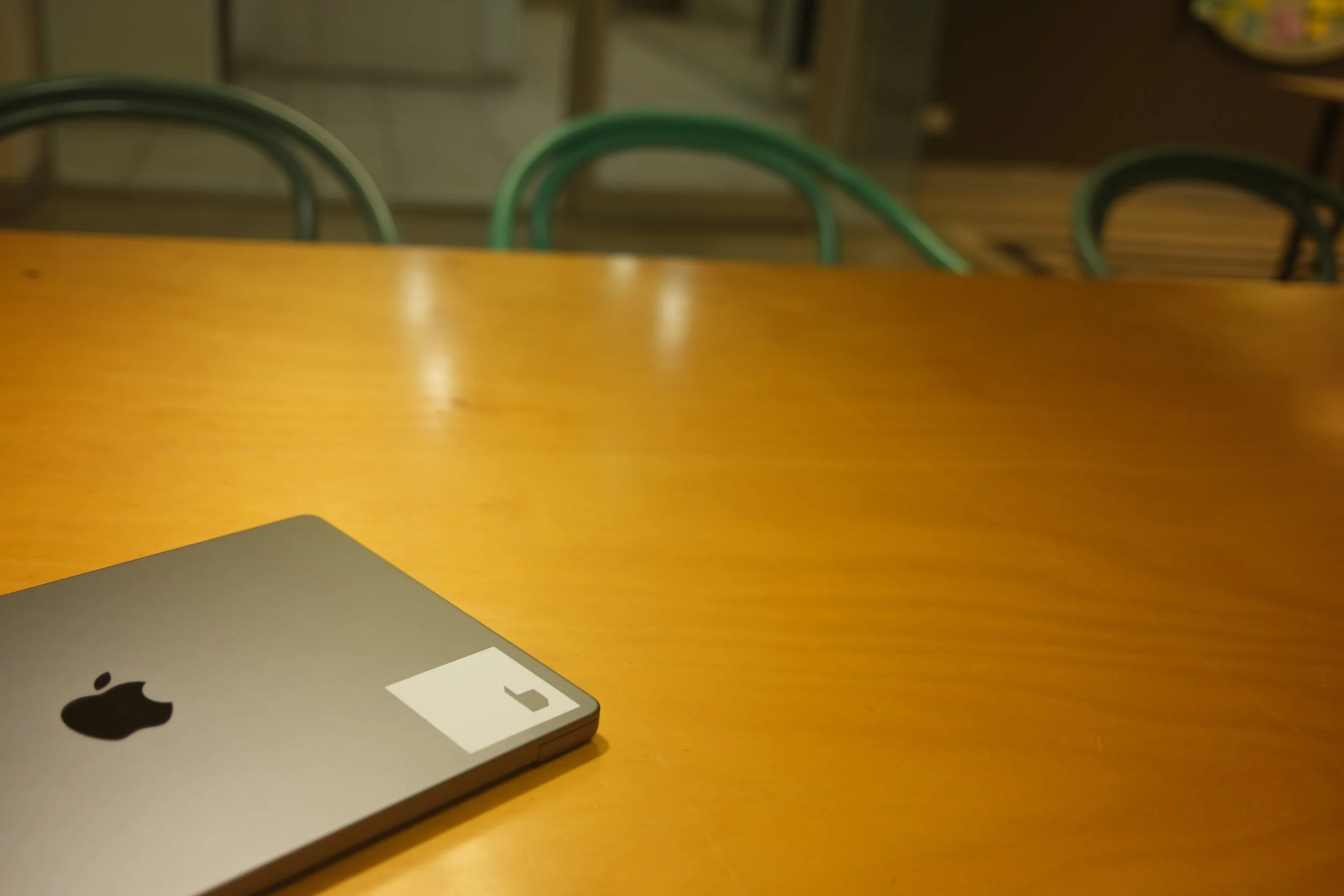 A closed silver MacBook laptop on a wooden table with an Apple logo and a paper sticker with the letter 'J'. In the background, there are metal chairs and a wooden shelf.
