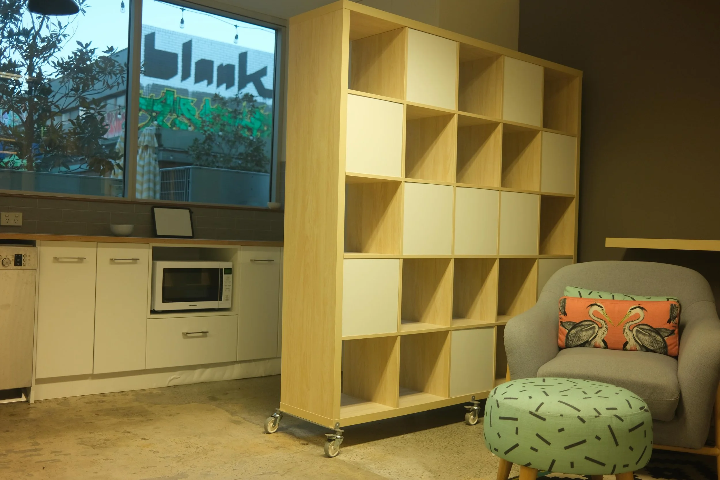 Interior of a modern kitchen with white cabinets, a microwave oven, and a window showing graffiti art outside. In the foreground, there is a gray armchair with a colorful flamingo pillow, a patterned ottoman, and a wooden rolling cart with empty shelves.
