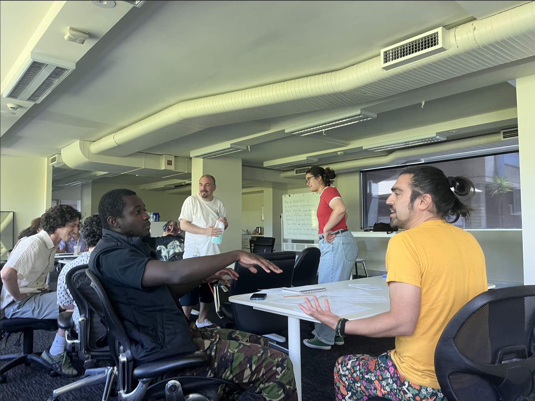 A diverse group of people participating in a workshop or discussion in an office conference room, with some sitting at tables and others standing near a whiteboard.