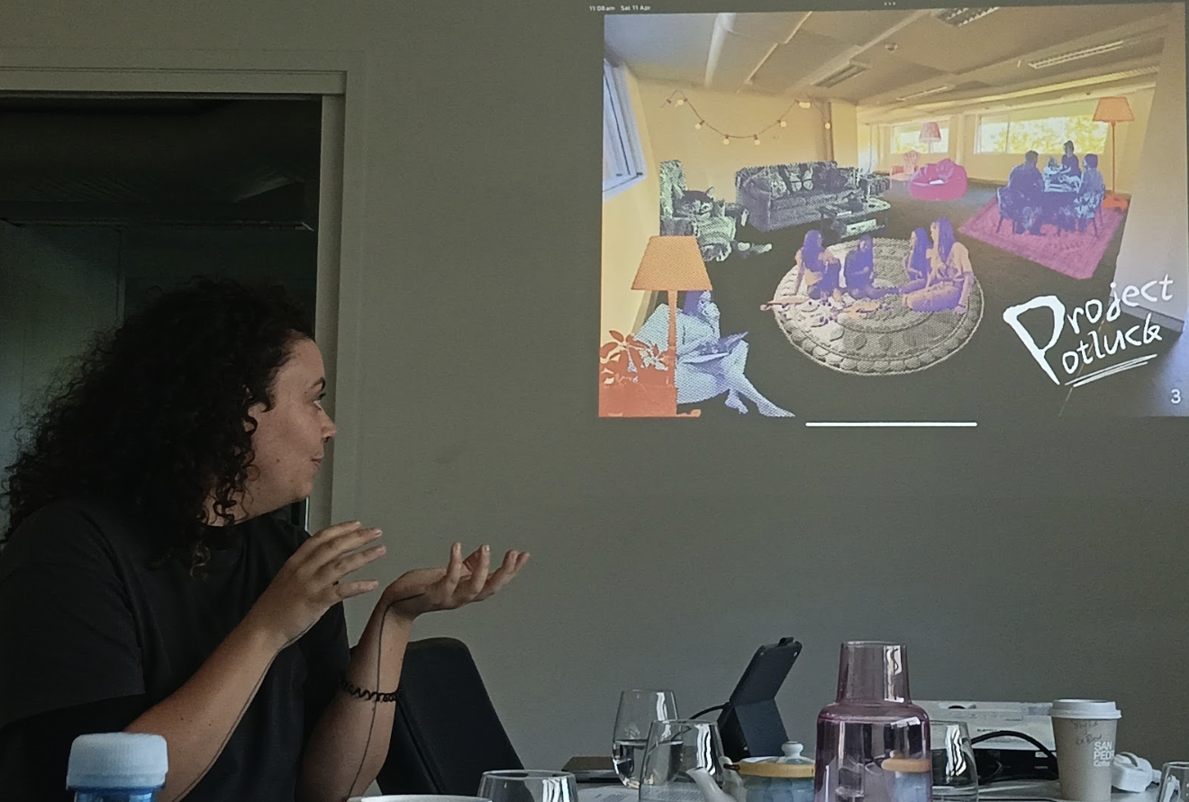 A woman with curly hair seated at a conference table, gesturing towards a projected slide on the wall. The slide displays an illustration of a cozy living room scene with several people engaging in various activities. The scene includes a sofa, armchair, coffee table, lamps, window, and string lights. The slide has the title 'Project Potluck' in the bottom right corner.