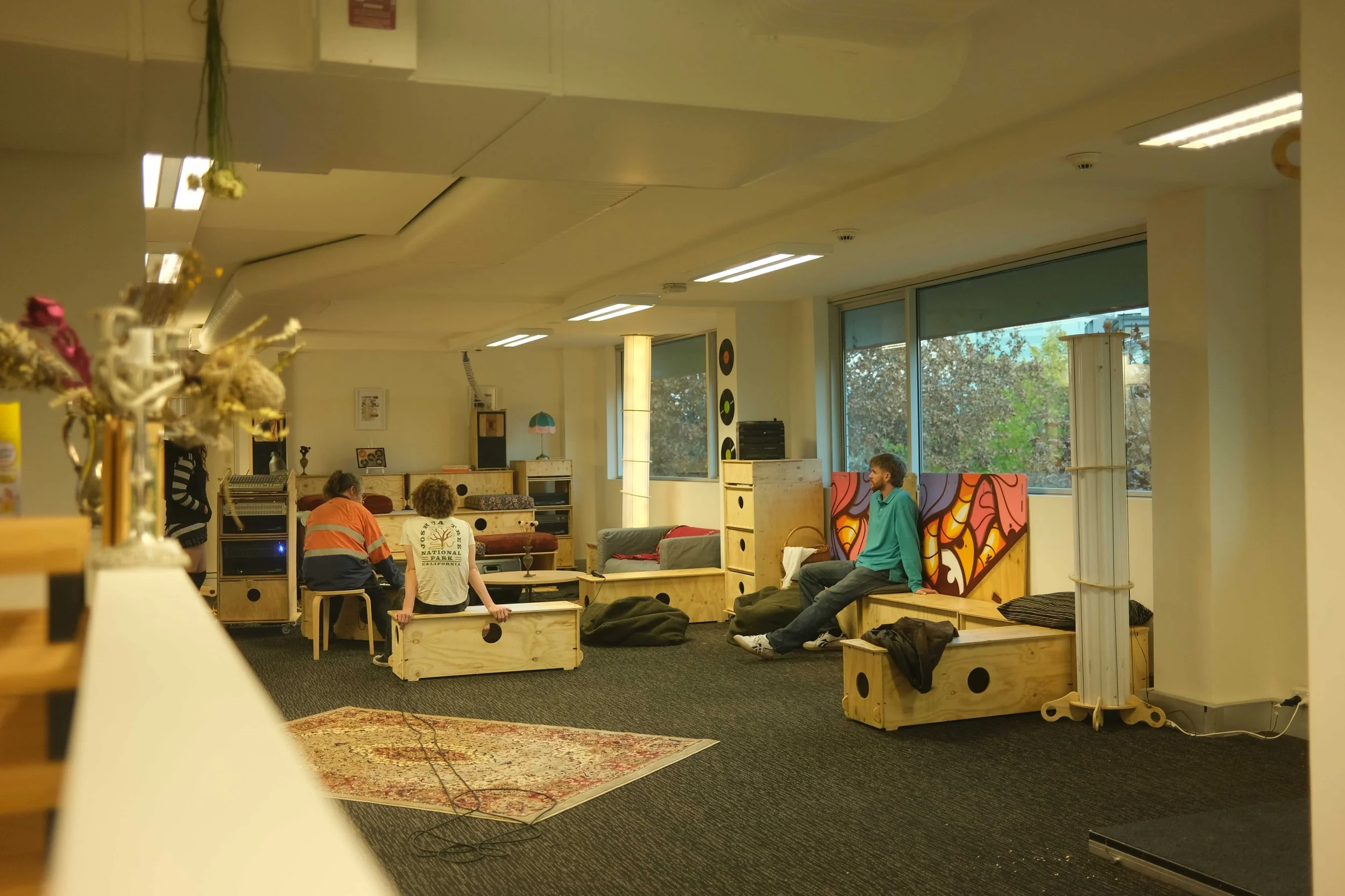 A group of people in a cozy, well-lit indoor space with large windows, sitting on wooden furniture and bean bags, engaging in conversation and relaxation; decorated with vibrant wall art and houseplants.
