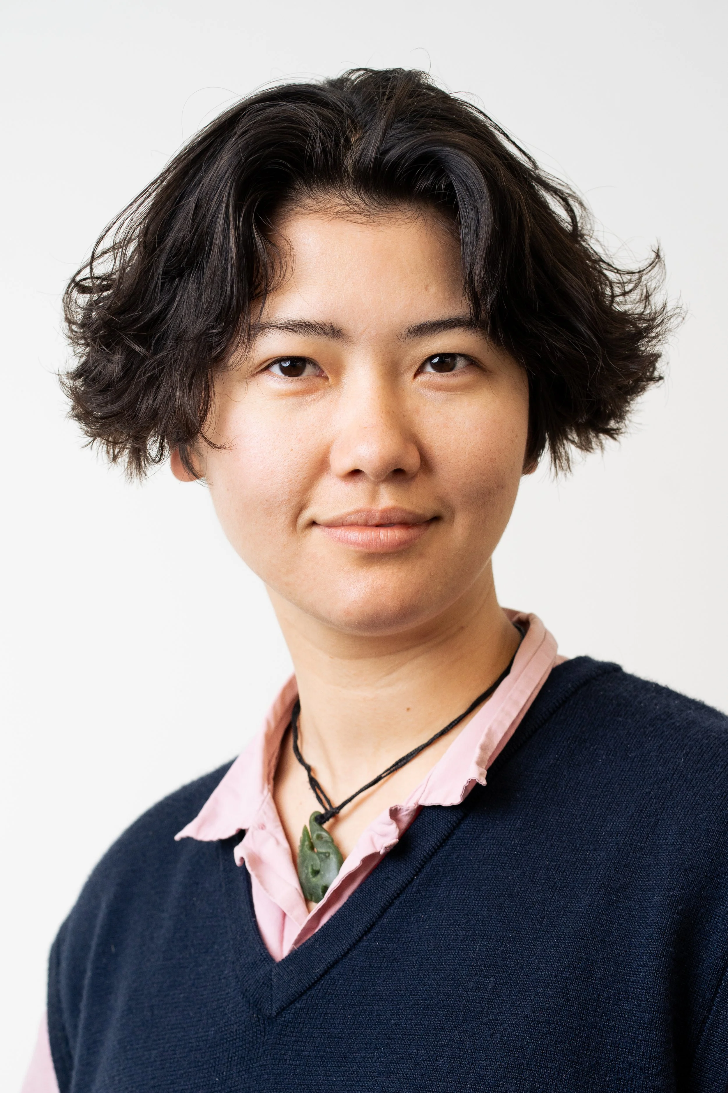 Close-up of a woman with short, wavy black hair, wearing a pink shirt with a black sweater over it, and layered necklaces, smiling subtly against a plain white background.
