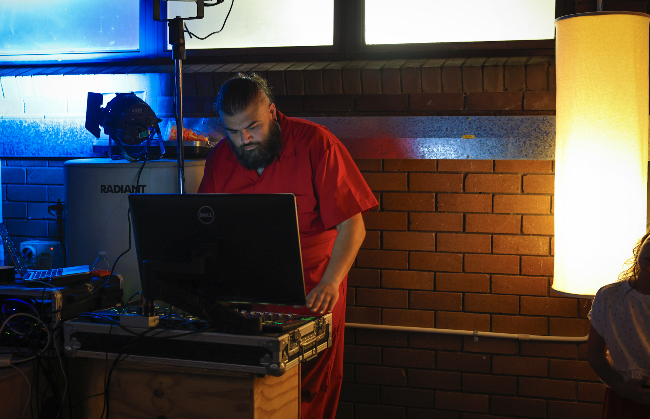 A man dressed in a red shirt is working with music equipment in a room with brick walls and a large yellow floor lamp. He appears to be focused on his work, with a laptop and electronic devices on a table in front of him.