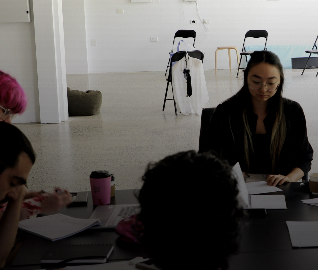 People sitting at a table with laptops, notebooks, and a pink reusable cup in a room with white walls and chairs in the background.