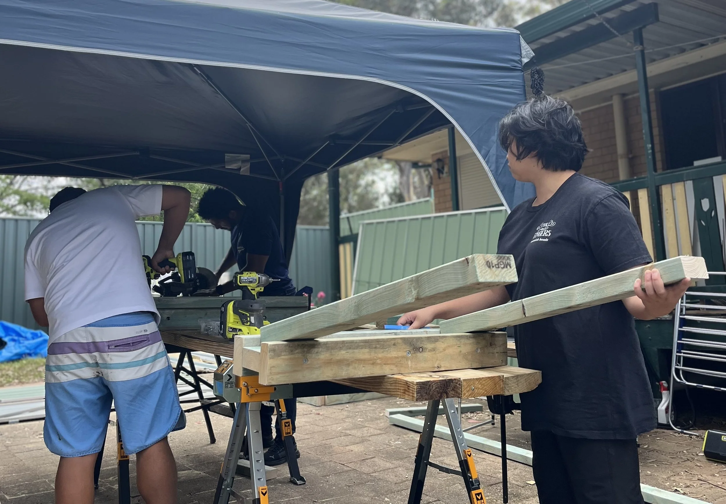 Children and adults working on wooden projects outside under a canopy, with tools and construction materials on tables.