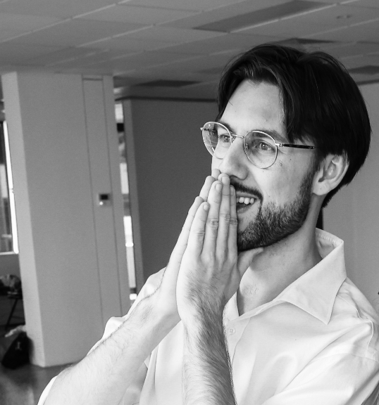 A black and white photo of a man with glasses and a beard, smiling and holding his hands over his mouth in surprise or amusement indoors.