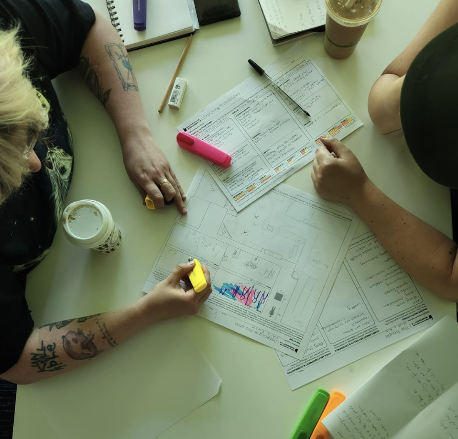 Two people working on a blueprint or floor plan on a white table, surrounded by notebooks, highlighters, a coffee cup, and an iced coffee.