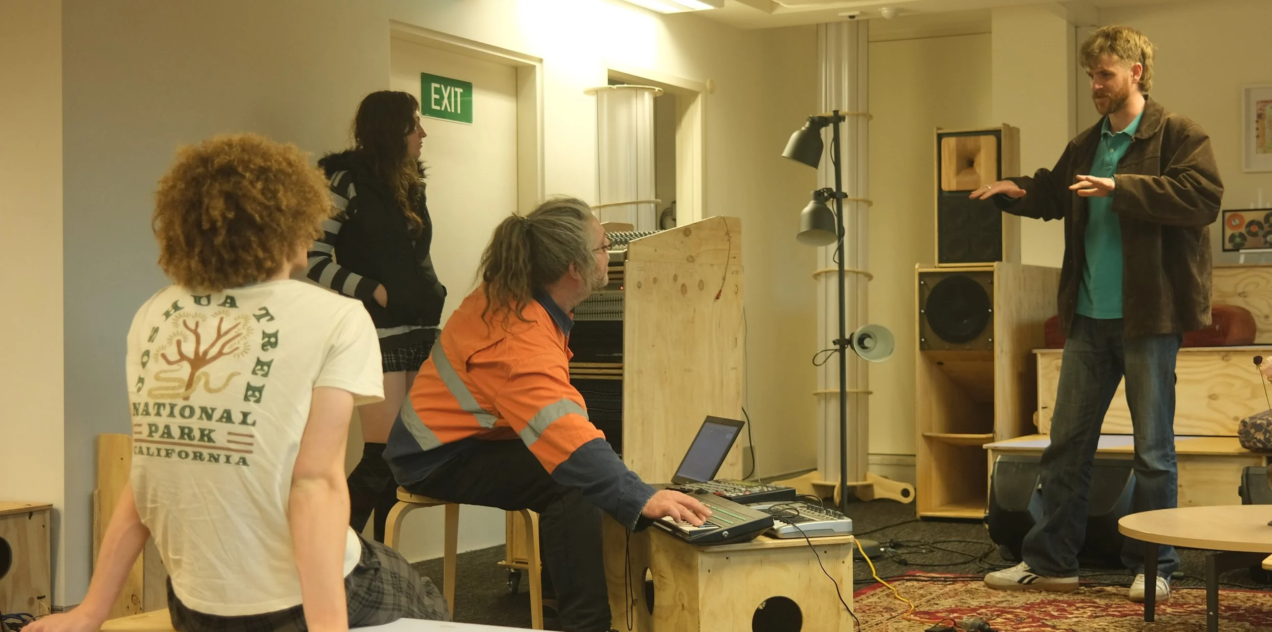 Group of four people in a room with musical and audio equipment. One man stands speaking, three others are seated, two at a table with electronic music gear, one woman standing. The room has bright lighting, wooden furniture, and a framed picture on the wall.
