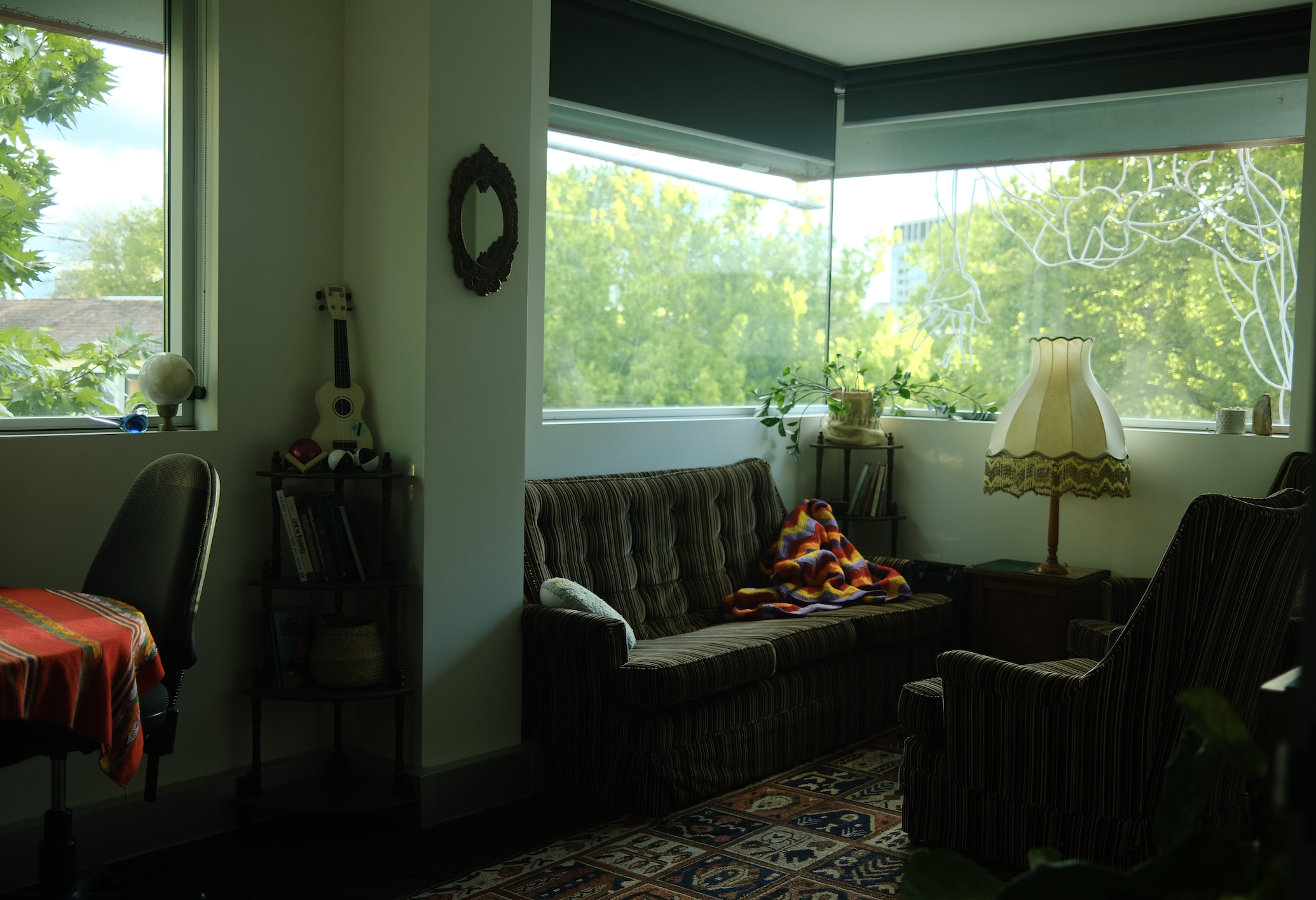 Interior of a living room with large windows showing green trees outside. There is a striped sofa with a colorful blanket, a vintage styled lamp, a small side table with plants, a framed mirror, a guitar on the wall, and a bookshelf.