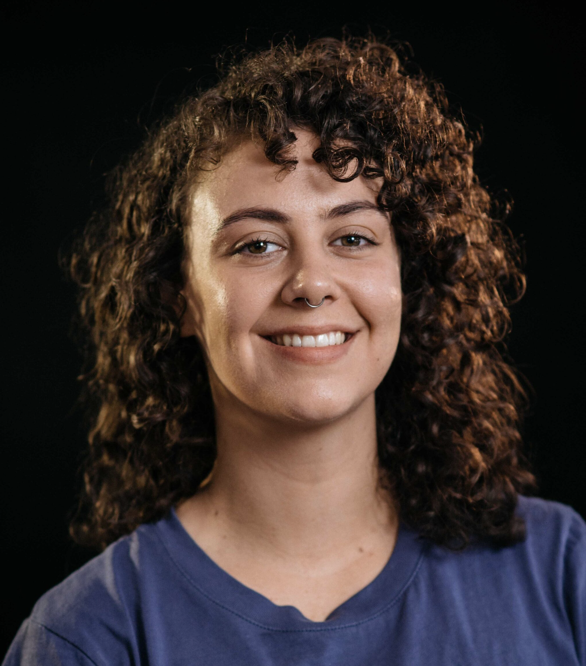 Portrait of a young woman with curly brown hair, smiling, wearing a blue shirt, against a black background.