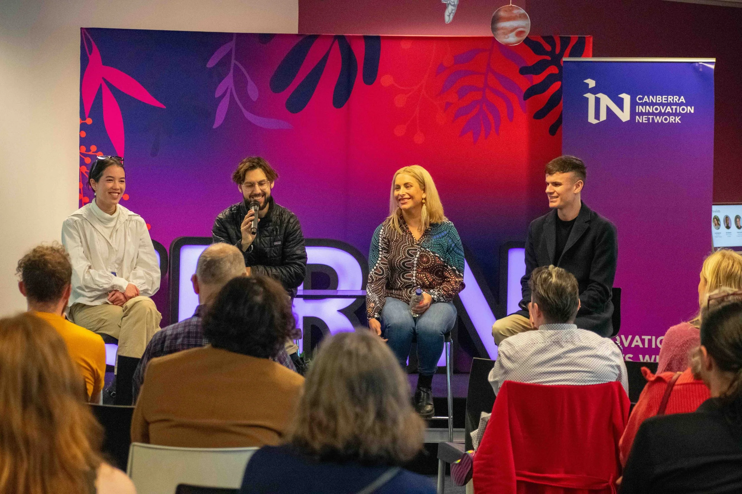 Four people are seated on a stage during a panel discussion or presentation, with an audience watching. The background features a purple and red design with the Canberra Innovation Network logo.