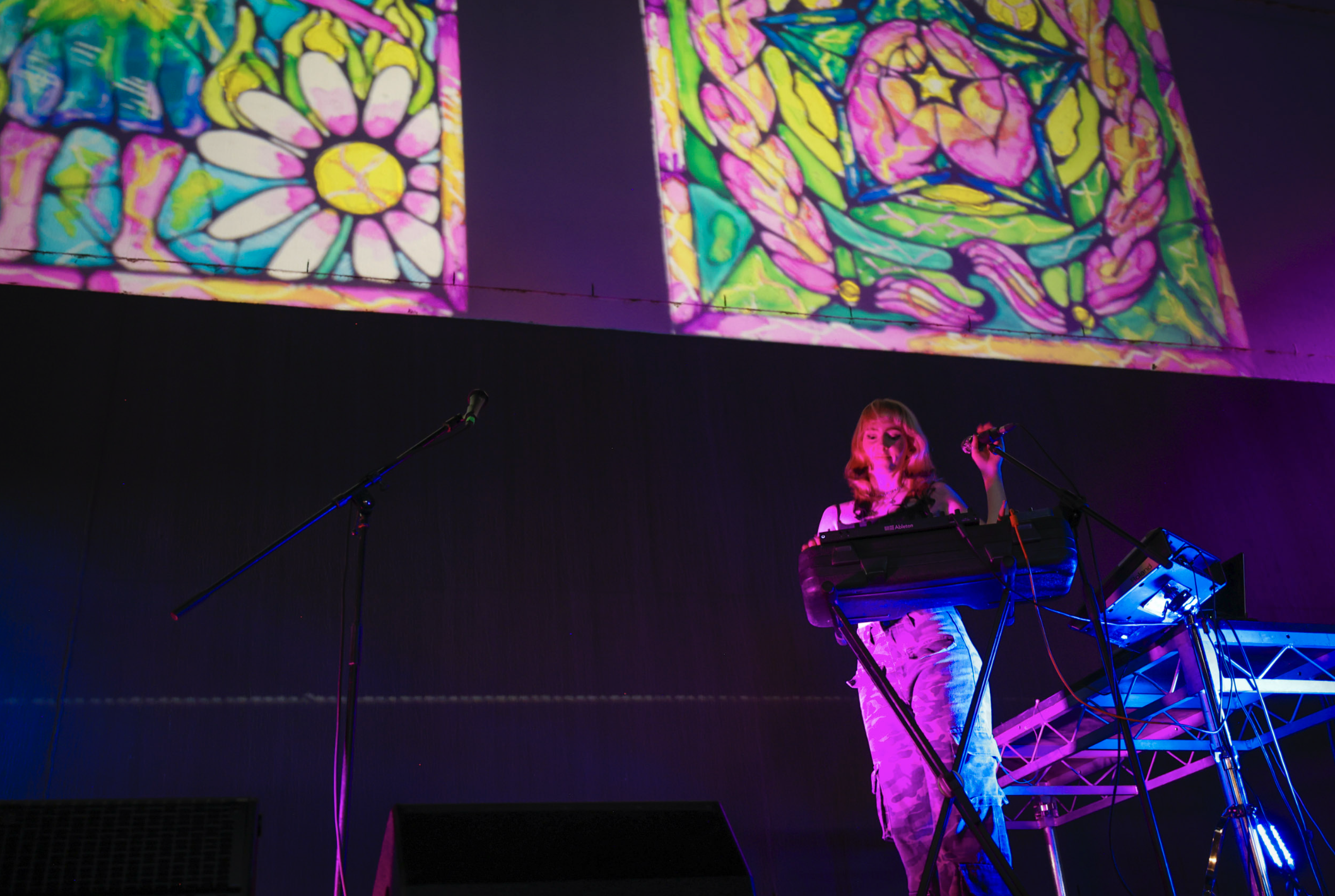 A woman performs on a stage with colorful stained-glass window projections behind her. She is playing a keyboard, illuminated by vibrant pink and blue stage lights.